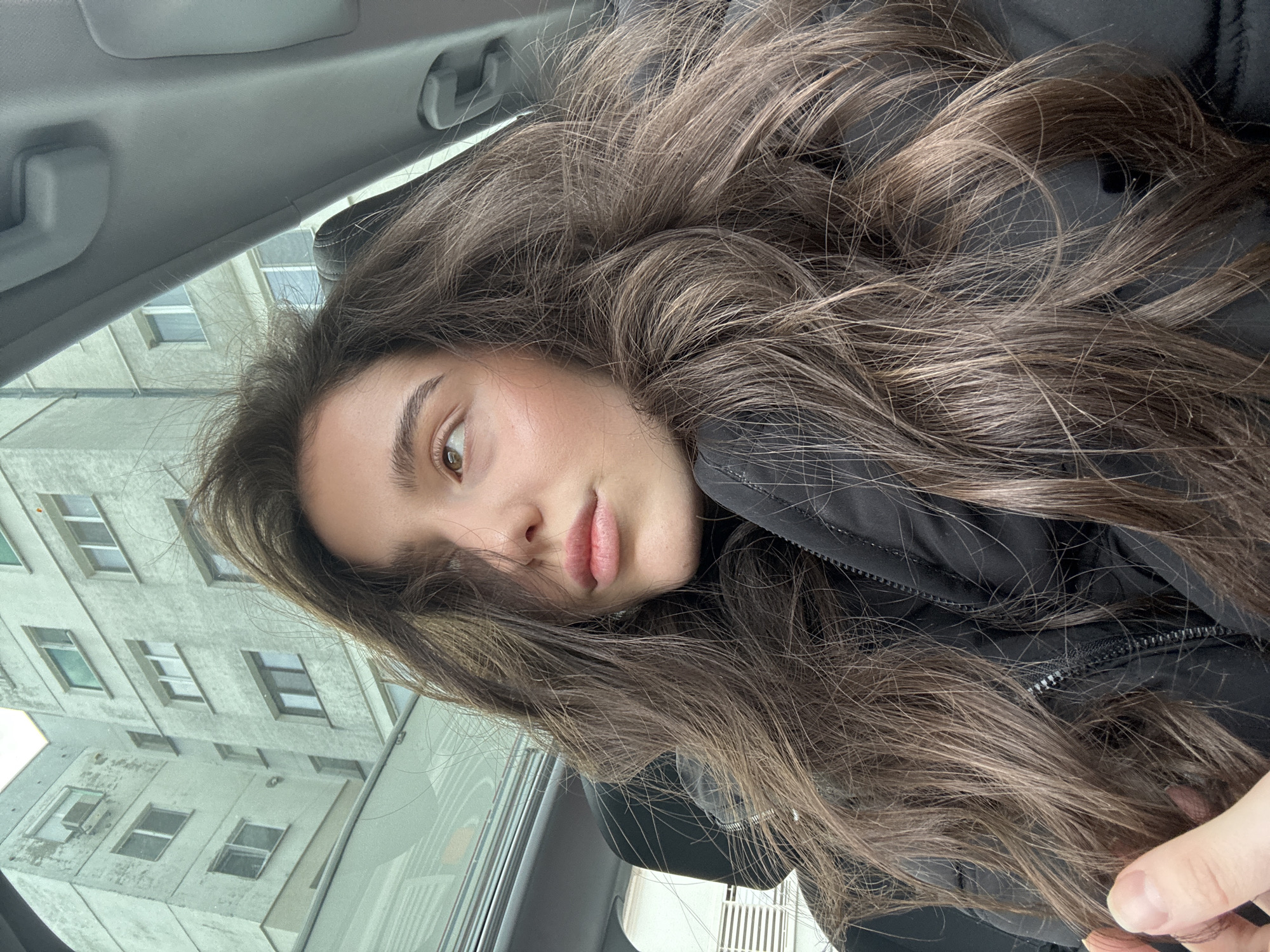 A young woman with long, wavy brown hair poses for a selfie. She is inside a car, evident from the car's interior visible in the background, which includes a gray headliner and a window frame. The building outside the car window is a multi-story concrete structure with several windows. She wears a black jacket with a visible zipper. Her skin tone is fair, and she has light brown eyes and full lips. The image appears to be taken at an angle, giving a slightly disoriented perspective. The location in the city of Unknown, Unknown, is unknown.