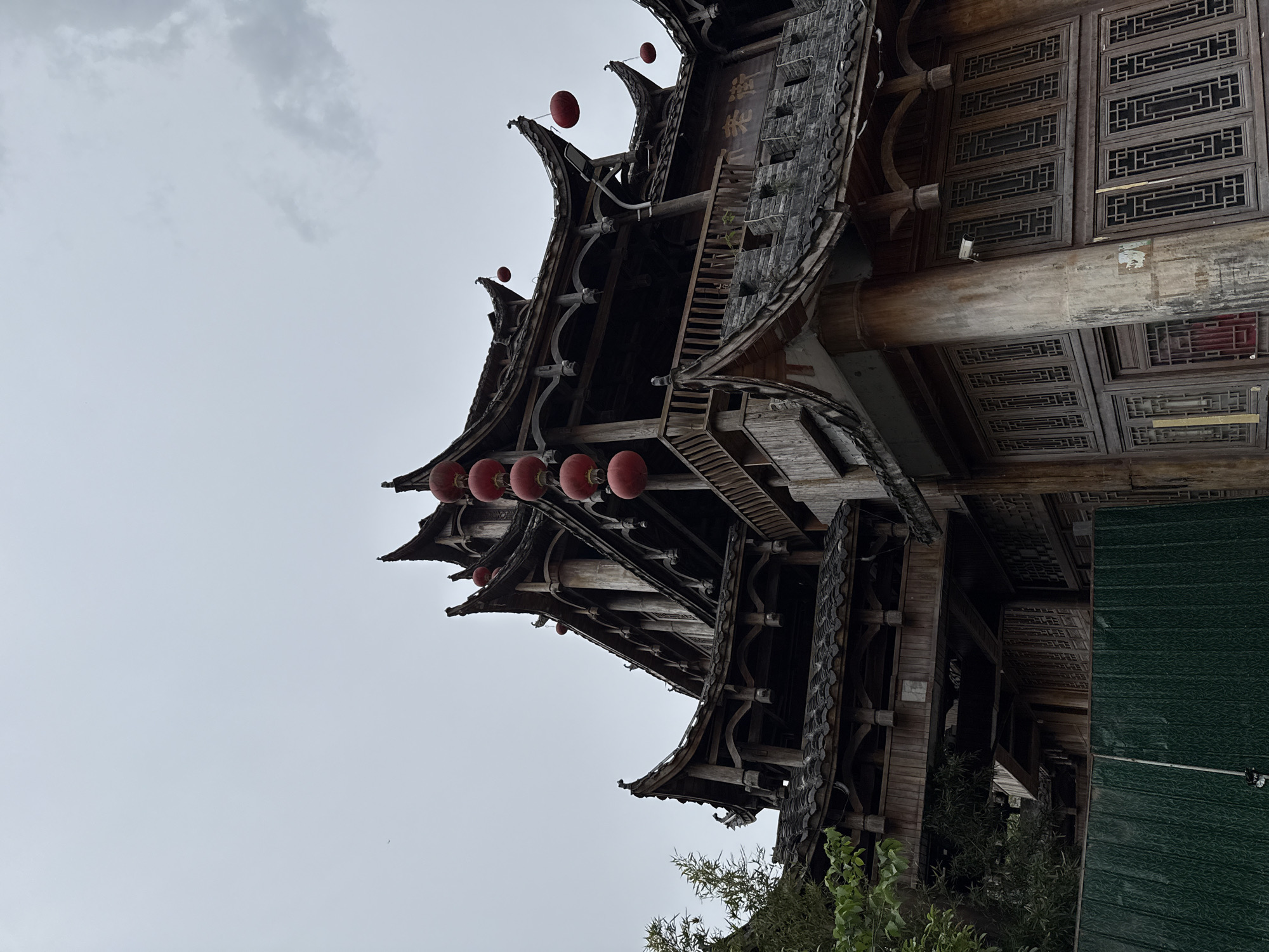 An upward-angle shot captures a multi-tiered traditional Chinese wooden building against a uniformly overcast, light gray sky. The building, likely a grand hall or a complex structure, features multiple layers of dark, intricately curved tiled roofs with upturned eaves. Red lanterns hang from various eaves and balconies, adding splashes of color to the otherwise muted palette of weathered dark wood and gray tiles.
The architecture is rich in detail, showcasing ornate wooden beams, pillars, and lattice-work panels on windows and railings. The wood appears aged, with visible textures and some wear, suggesting historical significance or traditional construction. A large, robust wooden pillar is visible supporting one section of the building. Further details include intricate geometric patterns on window frames and a security camera subtly placed on one of the wooden structures. Green foliage, possibly bamboo or other bushes, can be seen at the bottom of the frame, hinting at a natural setting around the structure.
On a vertical wooden plaque or sign near the top section of the building, Chinese characters are visible. The characters appear to read "濯溪山" (Zhuó Xī Shān), which translates to "Washing Stream Mountain" or "Washing Brook Mountain," possibly indicating the name of the establishment or a reference to its location. The scene depicts a quiet, possibly historical, moment under a cloudy sky in Wulingyuan, China, emphasizing the beauty and craftsmanship of traditional Chinese architecture. No specific activity or people are visible.