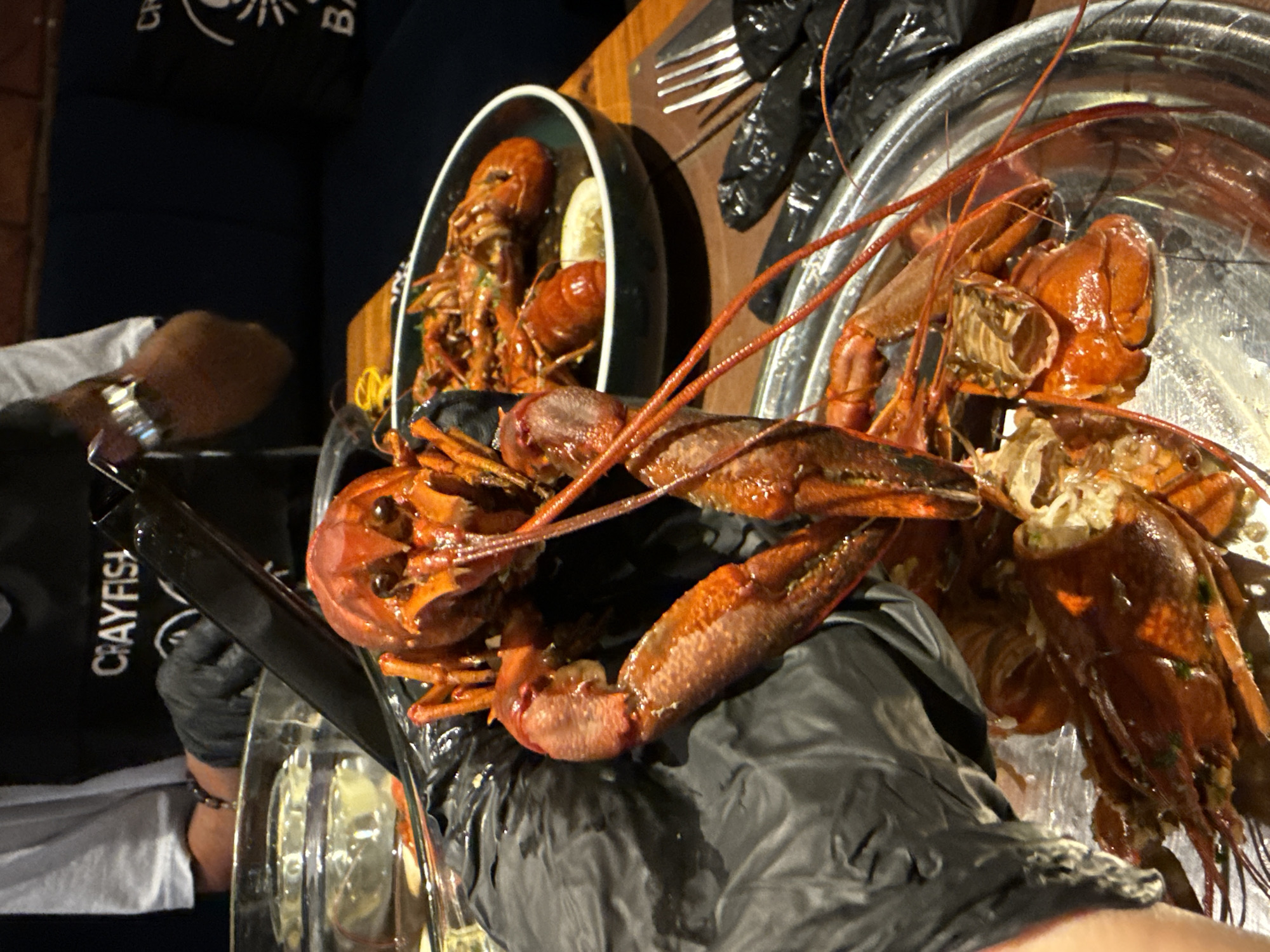 The image shows a selection of crawfish served on a table. There is a person wearing black gloves handling crawfish, with a partial view of their arm and wrist showing a bracelet and watch. Two types of containers hold the crawfish: a metal bowl and a black ceramic dish with a light-colored slice of citrus fruit. Cutlery and black plastic gloves are placed beside the dishes on a wooden table surface. The backdrop includes a dark blue couch and possibly a logo printed on a black shirt of the person holding the food. The location in the city of Kuta, Indonesia, is not indicated by any identifiable elements in the image.