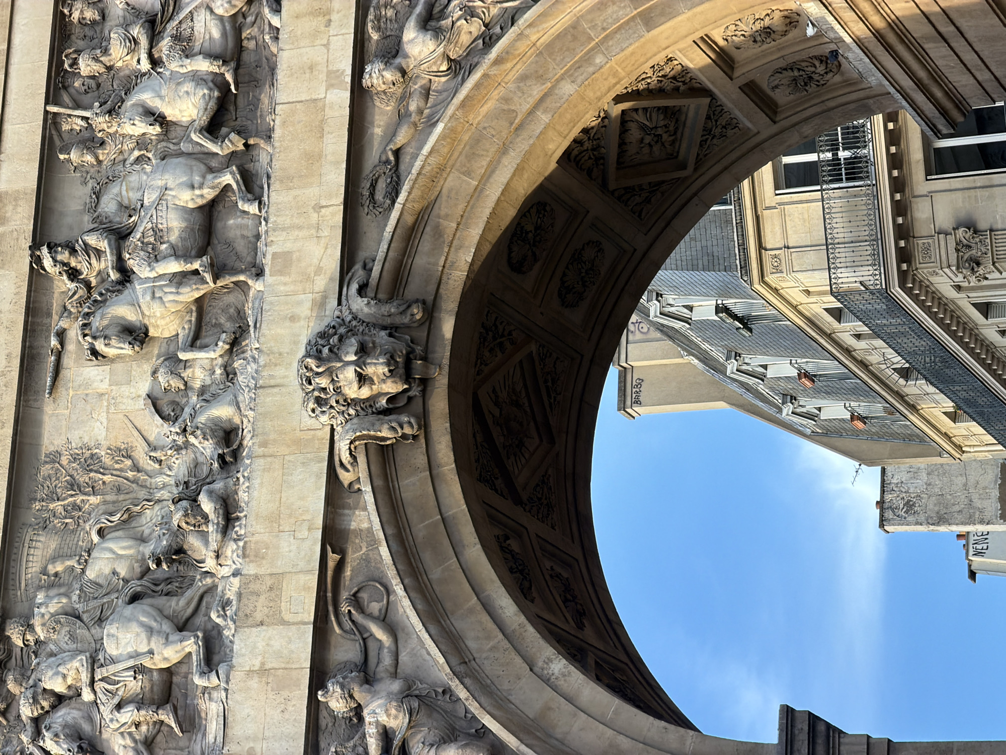The image displays a close-up, high-angle view of an ornate architectural structure, presented rotated 90 degrees counter-clockwise. The left portion of the frame is dominated by an intricate stone bas-relief running vertically. This detailed carving depicts a dynamic and dramatic scene, featuring multiple human figures engaged in battle or a struggle, intertwined with horses rearing, falling, and stumbling. The figures show considerable detail, suggesting warriors or mythological characters.
To the right, a grand stone archway spans diagonally across the composition. Its soffit (underside) is richly decorated with recessed coffers, each containing carved floral or abstract patterns. At the base of the arch on the left, a prominent sculpted head, possibly a keystone, features an elaborate headdress or flowing hair. Further along the arch's curve, additional sculptural elements are visible, including a figure seemingly holding a wreath.
Through the opening of the arch, sections of other buildings and a bright, clear blue sky are visible. The buildings exhibit typical Parisian architectural details, such as windows with dark frames and delicate iron balconies. The scene is illuminated by strong sunlight, casting distinct shadows that emphasize the depth and texture of the stone carvings.
On an upper edge of a building visible through the arch, graffiti spelling "BARBO" can be seen. Another partial inscription, "MENE", is visible on a different building section in the background. The overall impression conveys the historical grandeur and artistic craftsmanship of a major monument, consistent with structures found in Paris, France, under sunny conditions.