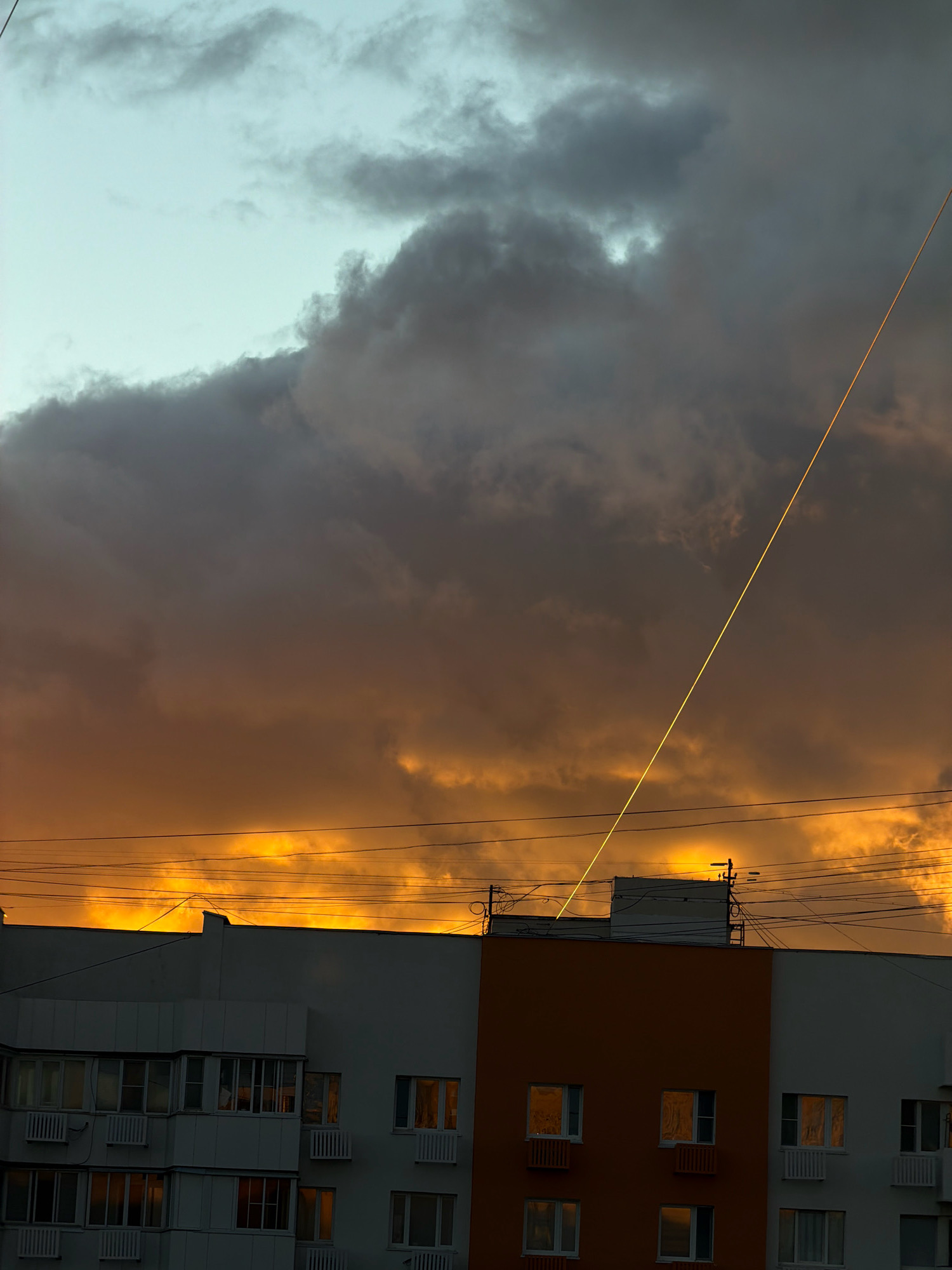 A wide shot captures the sky above a residential building in Khimki, Russia, during what appears to be sunset or sunrise. The sky is dominated by dramatic clouds, transitioning from a pale blue at the top to dark gray and then a vibrant, fiery orange near the horizon. A thin, bright yellow line, likely a contrail from an aircraft, cuts diagonally across the darker clouds. Below the sky, the upper portion of a multi-story apartment building is visible. The building has a mix of white and orange facades, with numerous windows reflecting the colors of the sky. Electrical wires are strung across the top of the building and into the frame. The overall scene is evocative of a stormy or dramatic atmospheric event occurring over an urban landscape. There are no people or distinct actions depicted in the image, only the static elements of the environment.