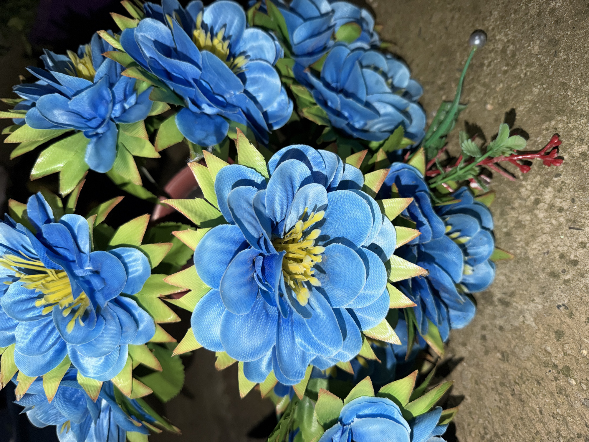 A close-up, high-angle shot captures a cluster of vibrant artificial blue flowers with bright yellow centers, set against a dark background and a weathered concrete or stone surface. The flowers, crafted from fabric-like material, feature multiple layers of petals in varying shades of blue, creating depth. Each flower is surrounded by pointed, light green leaves, possibly made of plastic, some with a subtle yellow or brown edge.
In the upper right portion of the image, emerging from the dark background, a single slender green wire stem topped with a small, reflective silver bead is visible. Below it, a small sprig of artificial red berries or buds on a green plastic branch adds a contrasting color element to the arrangement.
The concrete or stone background, visible on the right and bottom right, appears textured and somewhat aged, suggesting an outdoor setting or a durable surface. The lighting creates distinct shadows beneath the flowers and leaves, indicating a specific light source, possibly artificial or directional sunlight. The overall arrangement suggests a decorative display, potentially for a memorial or grave, consistent with the location context of Dabene, Bulgaria. No text is visible in the image.