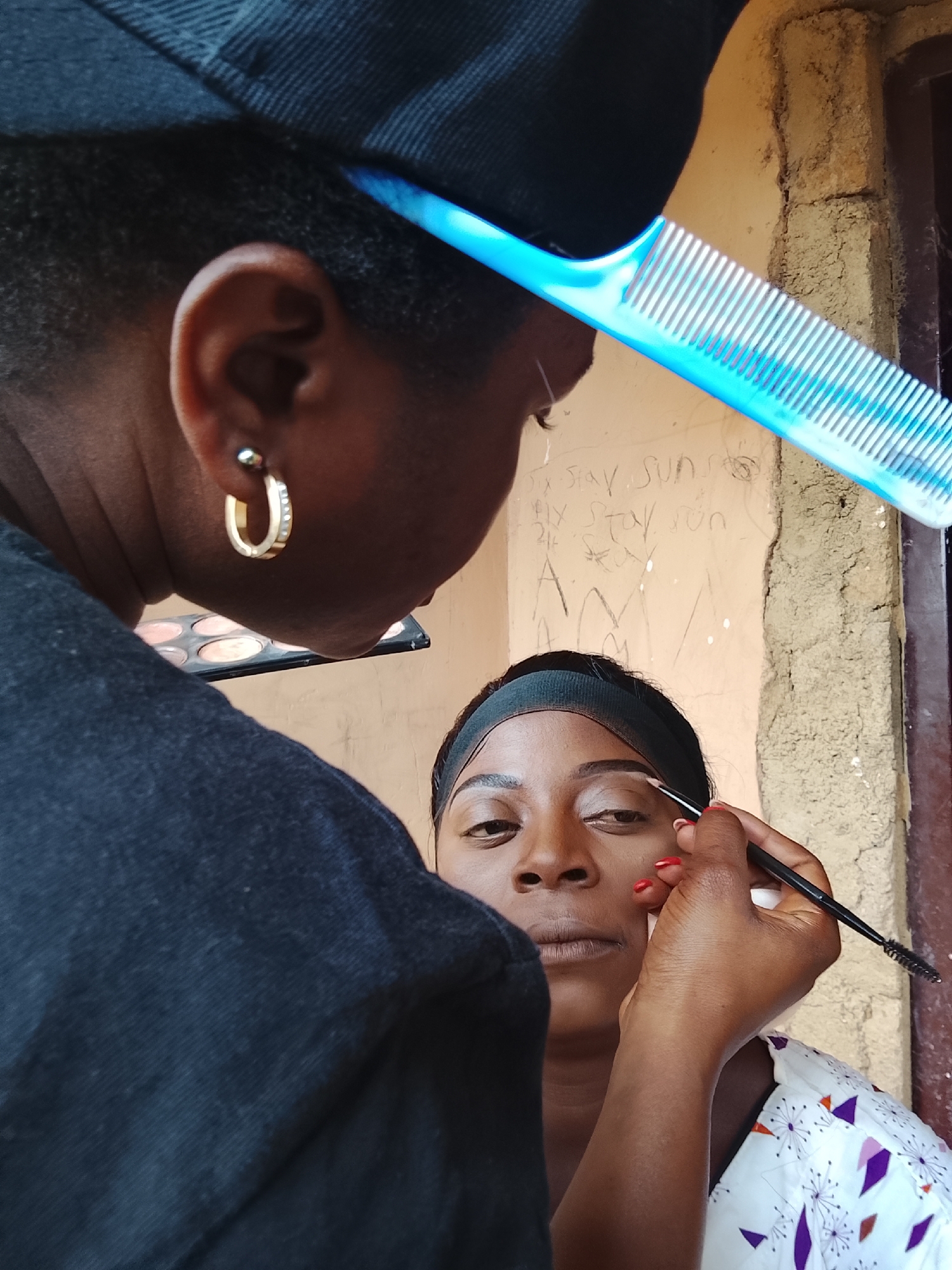 A photograph taken in an unknown location in Unknown, Unknown, depicts a woman receiving eyebrow makeup application. The makeup artist, positioned on the left, is wearing a black shirt and a black baseball cap with a blue comb placed on her head. Her left ear is visible, adorned with a gold hoop earring. She is holding a makeup palette. The client, facing forward, has a black headband on her head and is wearing a white shirt patterned with purple and orange shapes. The makeup artist is using a black makeup brush to apply product to the client's eyebrow. The background consists of a textured wall with graffiti.