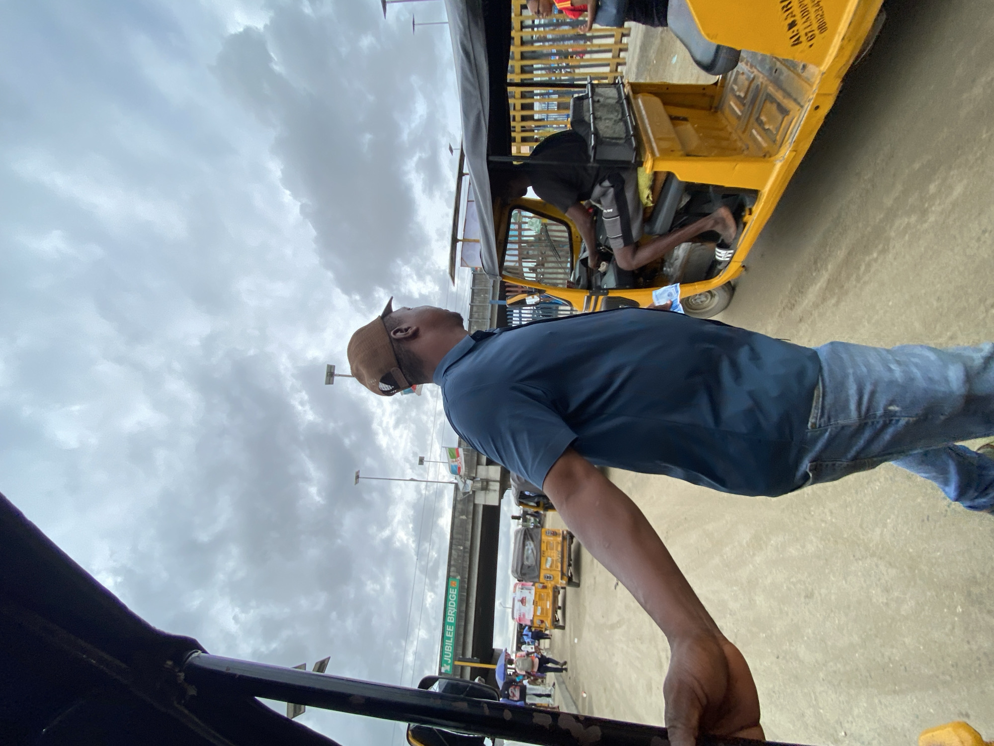 The image captures an outdoor urban scene in Lagos, Nigeria, under an overcast sky. In the foreground, a man is seen in profile, wearing a blue short-sleeved shirt, blue jeans, and a light brown cap. He stands with his left hand resting on a black railing, looking upwards.
In the middle ground, a bright yellow commercial tricycle, characteristic of Nigerian transportation (Keke Napep), is visible. A person in a dark shirt and shorts is seated inside, with bare feet. On the seat, small bundles that appear to be money or goods are present. The side of this tricycle has partially visible text, including "AL. M. RA..." and a numerical sequence beginning "080234...".
The background features a large concrete overpass or bridge. A prominent green sign on the bridge clearly displays the text "JUBILEE BRIDGE". Beneath the bridge, other people are visible, some walking, and another yellow tricycle can be seen further down the street. One individual in the distance is holding a blue umbrella. The ground is a light-colored paved surface, suggesting a street or market area. The sky above is entirely covered with thick, grey clouds, indicating an overcast day. The scene depicts a typical, busy urban environment.