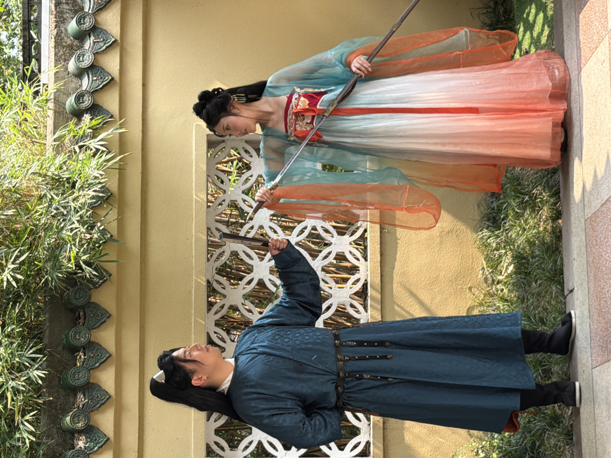 In an outdoor setting within Yuzhong District, China, two individuals in traditional East Asian attire are positioned opposite each other. The male on the left, with black hair pulled into a ponytail secured by a silver hairpin, wears a dark teal, patterned robe with a white collar and a dark belt adorned with metal studs and hanging straps. He holds a closed black folding fan in his right hand, extending it forward. The female on the right, featuring an elaborate black updo with golden hair ornaments, wears a multi-layered dress: a red embroidered top, a white-to-orange gradient skirt, and a sheer teal outer robe with orange sleeves. She holds a long, silver-colored staff or sword, pointing its tip towards the male's extended fan with both hands. Both subjects are looking directly at each other. The background consists of a light yellow textured wall featuring a white decorative lattice screen. Above the wall, a row of green, ornate Chinese roof tiles is visible, surmounted by dense green bamboo foliage. The ground is paved with checkered tiles in varying shades, flanked by patches of green grass and small plants.