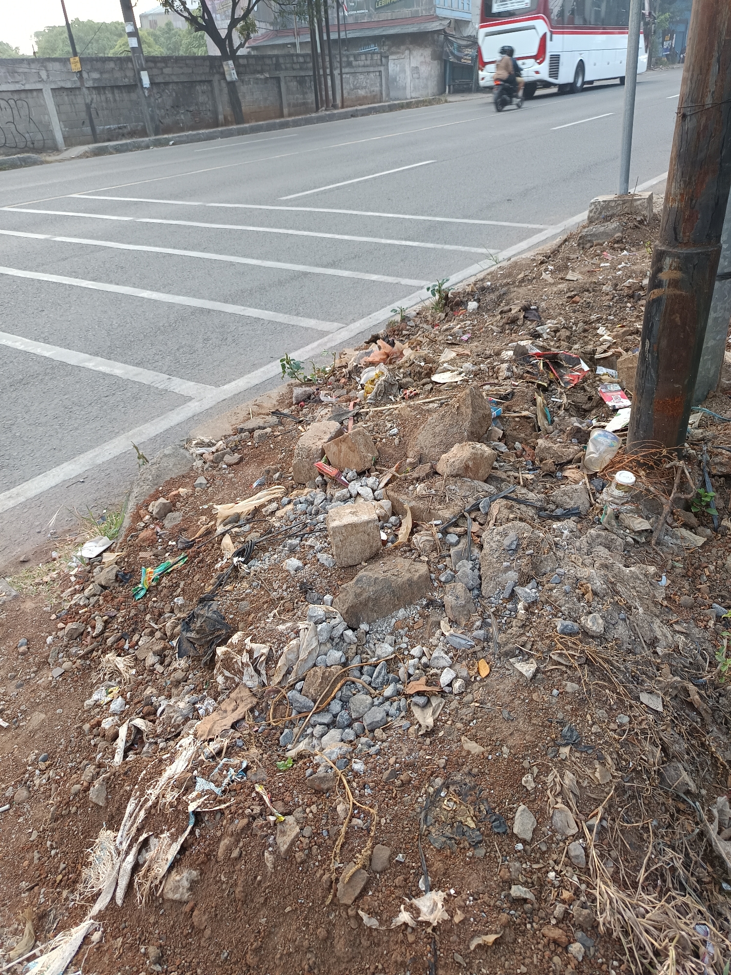 In Talaga Sari, Indonesia, a roadside scene is captured, focusing on a section of debris-strewn earth bordering a paved road. The road features distinct white lane markings and a pedestrian crossing. A white bus with red trim travels on the road, accompanied by a motorcyclist. The roadside area is densely covered with various types of trash, including plastic bags, crumpled paper, rocks, and unidentifiable waste. A rusty metal pole stands vertically within the frame, adjacent to the accumulated refuse. The ground is a mix of brown soil and small stones, with sparse vegetation poking through. The overall impression is one of urban neglect and litter.