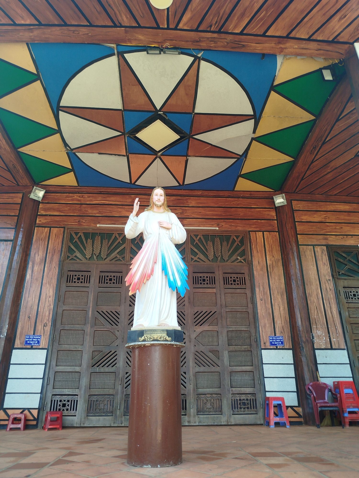 The image shows a statue of Jesus Christ under a decorated ceiling in An Phu Ward, Vietnam. Jesus stands on a brown cylindrical pedestal, wearing a white robe with red and blue emanations from his heart. He is depicted with his right hand raised in blessing and his left hand near his chest. The background features ornate wooden doors and panels. Above, a geometric ceiling design incorporates blue, green, and brown triangles and shapes. Two small blue signs are affixed on the left and right side of the door with Vietnamese writing. Small red chairs are placed in front of the doorway. The tile flooring appears to be terracotta-colored. Two exterior lights are visible.