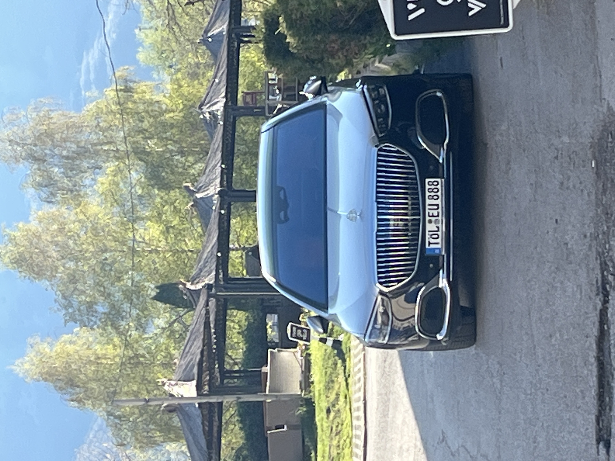 The image shows the front of a white and black car parked on a street. The car has a license plate that reads "TOL EU 888". The background includes trees, a structure with a black roof, and a street. The location appears to be in Sofia, Bulgaria, based on the "TOL" license plate code for the Tólna county in Hungary. The car appears to be parked, and there are no visible people interacting with it. The environment seems to be a street lined with trees and some structures.