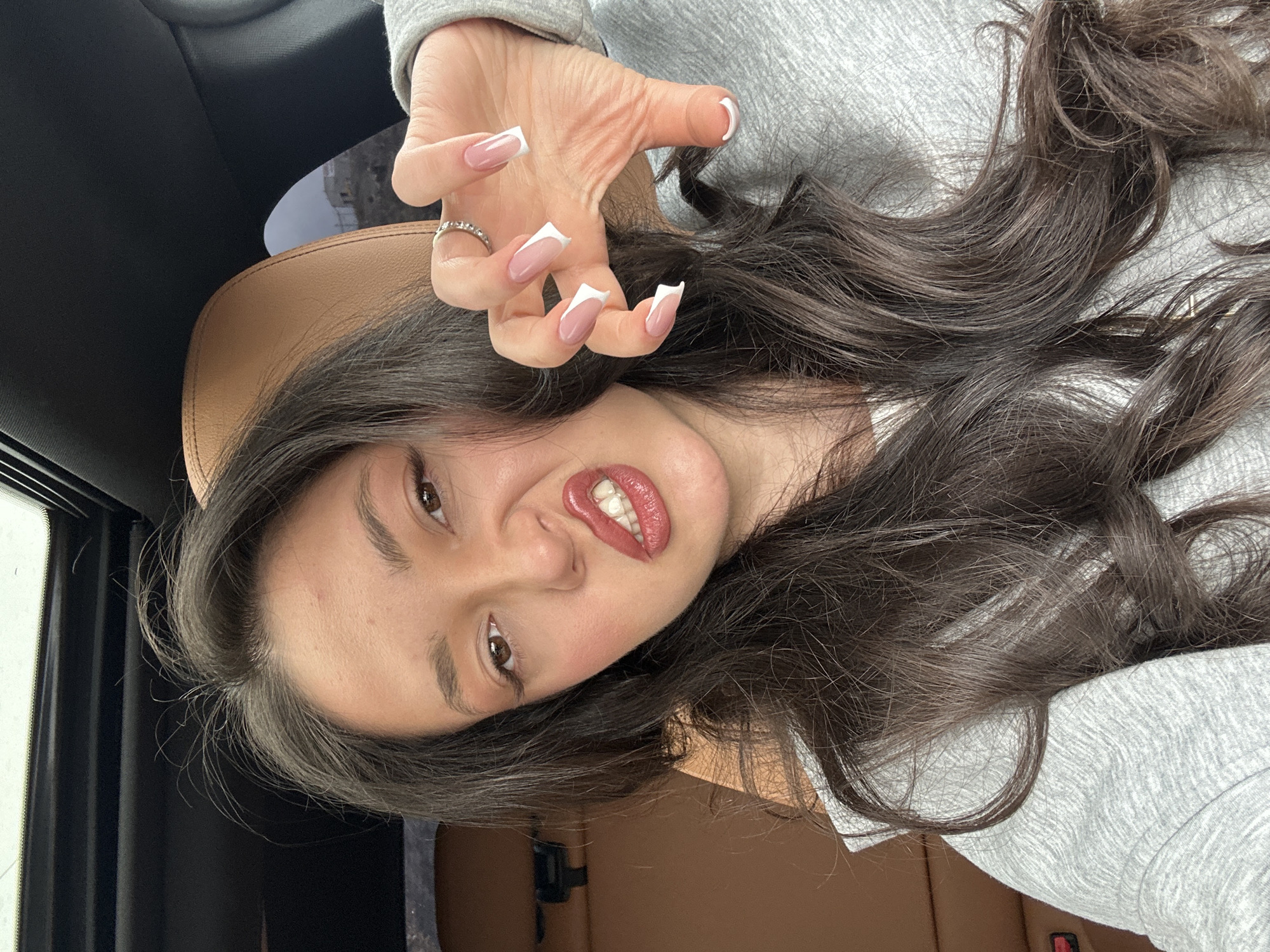 The image presents a close-up portrait of an adult female individual seated in the front passenger seat of a vehicle in Yerevan, Armenia. The subject has long, dark brown hair and is wearing a light grey hooded sweatshirt. Her face is directed towards the camera, exhibiting a playful contortion: her upper lip is curled to expose her upper teeth, her mouth is slightly open, and her brow is furrowed. Her right hand is raised towards the camera, fingers splayed and bent in a deliberate, claw-like gesture. Her nails are long, shaped with squared tips, and feature a white-tipped French manicure design. A silver-colored ring with small stones is visible on her right ring finger. The vehicle's interior includes a brown leather headrest behind the subject and a black roof lining with a visible sunroof. Through the car window, a distant, light-colored hillside with an indistinct structure and fencing is observed under an overcast sky.