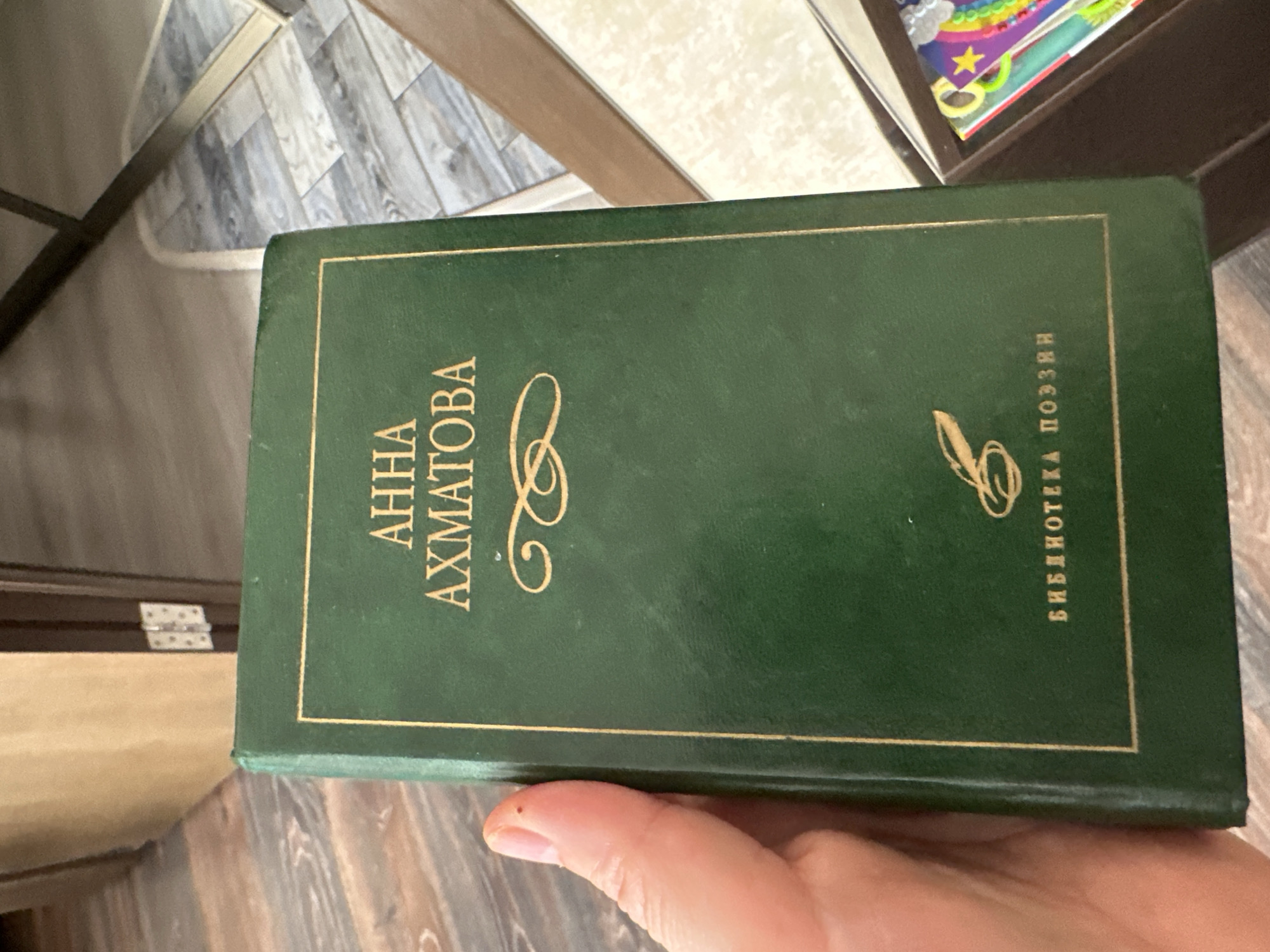 An open green book with gold lettering and trim is held in a person's left hand. The book's cover displays the name "АННА АХМАТОВА" in gold capital letters, with decorative flourishes and a quill pen graphic above the words "БИБЛИОТЕКА ПОЭЗИИ". The background shows a wooden floor and a partially visible doorway with a hinge. The environment appears to be indoors, likely a residential or personal space, and not in the city of Podolsk, Russia.