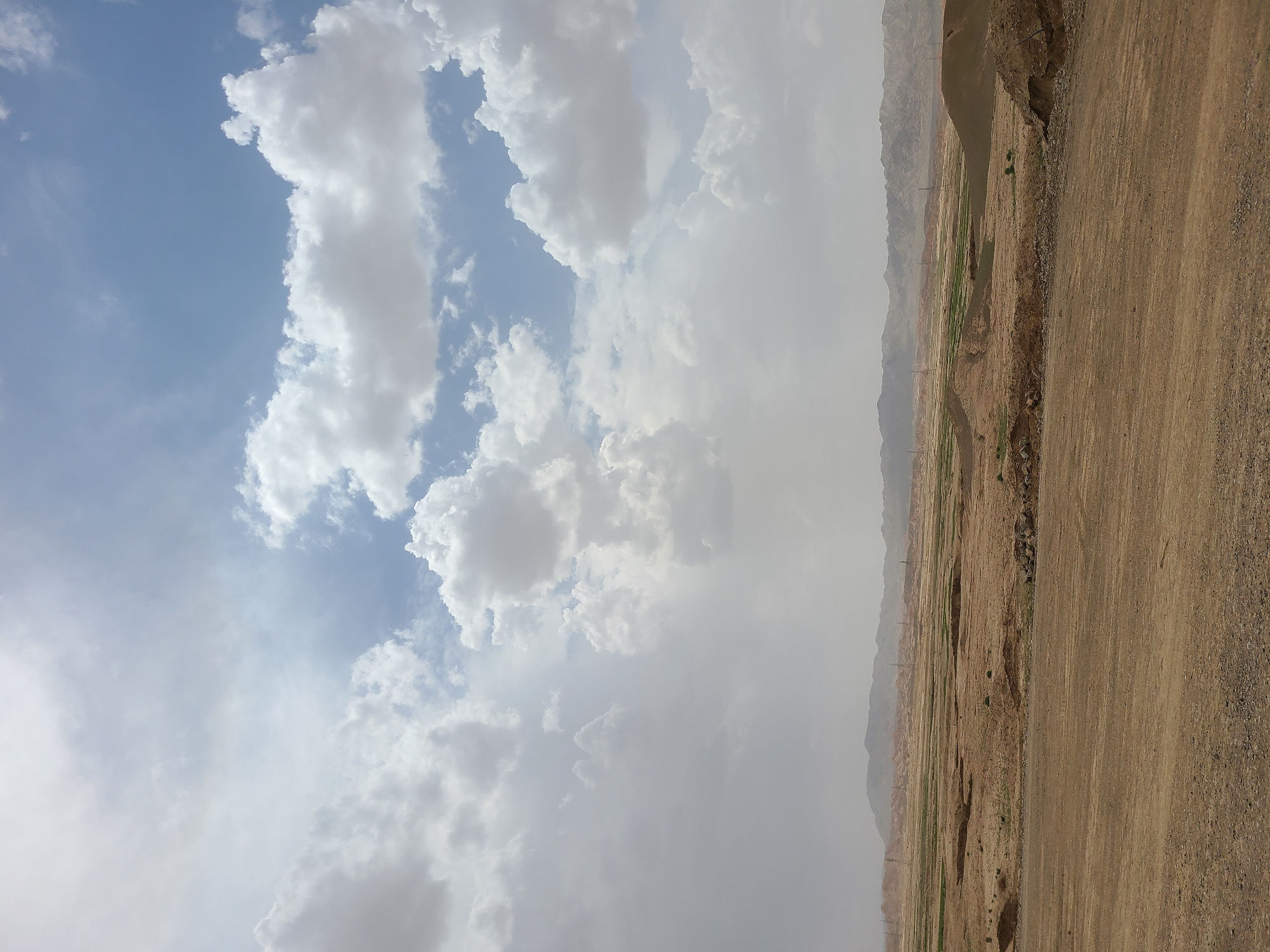 This image captures a wide outdoor landscape under a partly cloudy sky, characteristic of an arid or semi-arid region. The sky dominates the upper portion of the frame, featuring a mix of light blue patches and numerous large, fluffy cumulus clouds that vary in color from bright white to light grey. A general atmospheric haze is noticeable, especially towards the horizon.
The lower third of the image presents a dry, expansive terrain. In the foreground, a broad, unpaved dirt road or track stretches across the right side. Beyond this track, the midground consists of undulating, brown earth with sparse patches of green vegetation, suggesting some limited cultivation or natural growth in depressions. Several thin, dark utility poles are visible stretching into the distance. In the background, a range of hazy mountains or hills lines the horizon, their muted colors blending into the atmospheric haze.
No people or animals are visible, and no specific activity is taking place; it's a static landscape view. The time of day appears to be daylight, possibly mid-morning or afternoon, given the bright illumination filtered through the clouds. The scene conveys a sense of vastness and natural tranquility. There is no visible text in the image. The environment is consistent with the geographical characteristics of Kuchlak, Pakistan.
