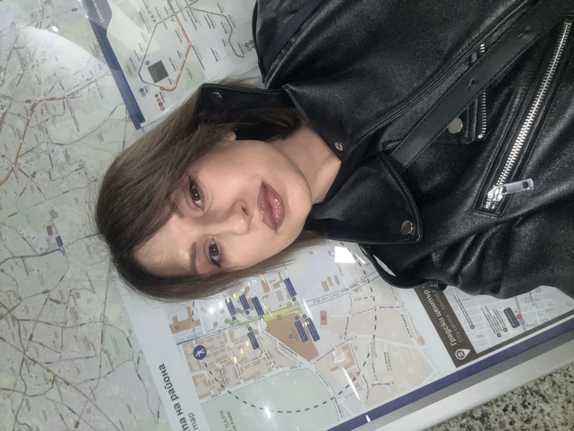 A woman wearing a black leather jacket is lying on top of a city map. The map appears to be a public transportation or tourist map. Text visible on the map includes "Градски център City centre - tourist", "Схема на района map", "Парк", "бул. Черни връх Cherni vrah Blvd", and "Ген. Иван Колев". The map appears to depict a portion of Sofia, Bulgaria.
