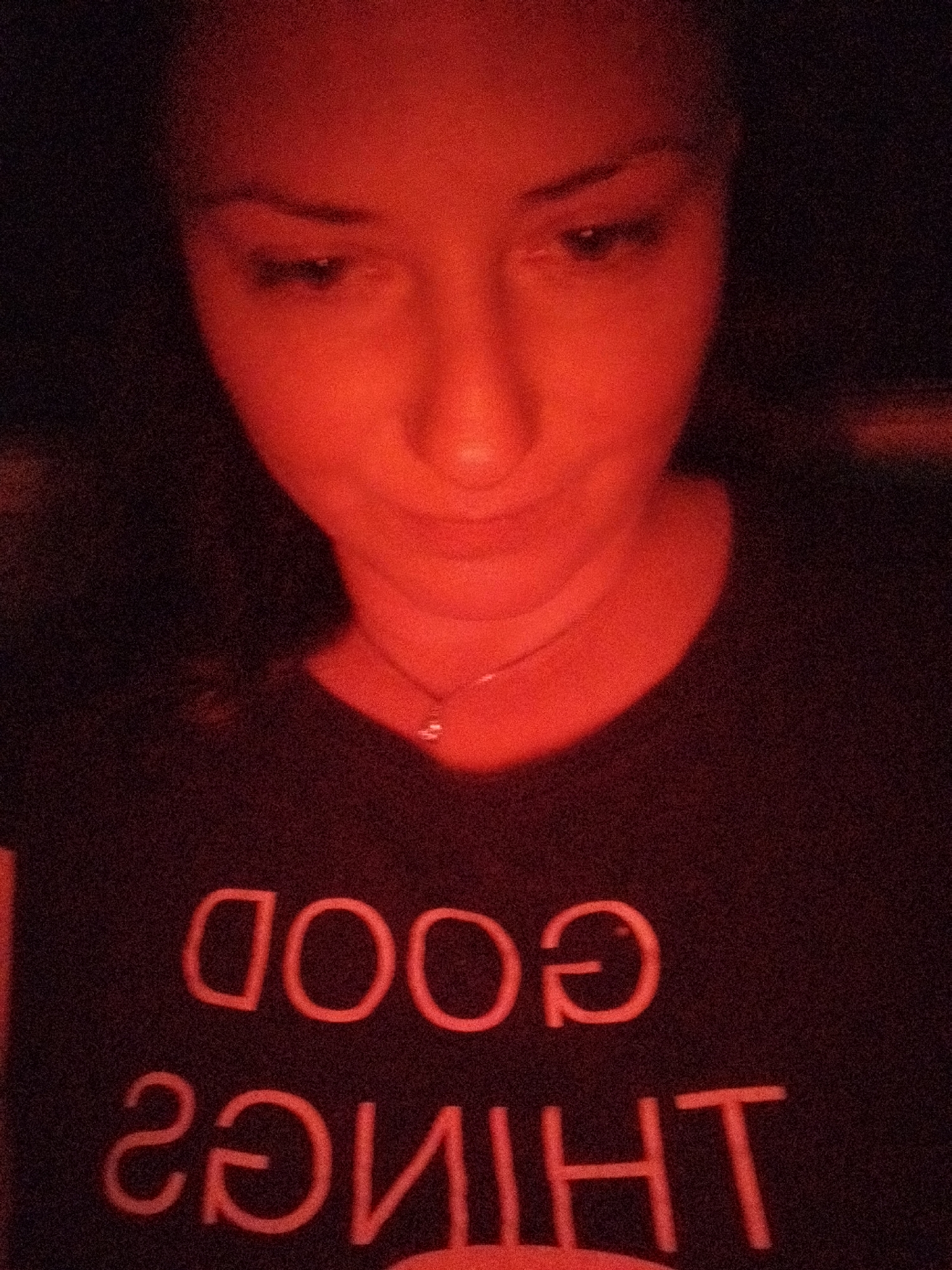A woman is shown indoors, bathed in a strong red light, possibly from a lamp or lightbulb. She has fair skin and dark hair and is wearing a dark t-shirt with the reversed text "GOOD THINGS" printed on it. She has a delicate necklace around her neck. The background is very dark, making it impossible to discern anything about the location. No evidence in the image to suggest the location is Sofia, Bulgaria.