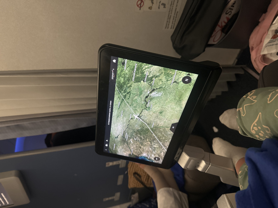An airplane in-flight entertainment screen displays a satellite image map. The plane's current location is marked. Passengers are visible in the frame, including feet and legs. Overhead compartment, airplane windows, and seat details are also visible. The screen shows a navigation app, potentially tracking the flight path.