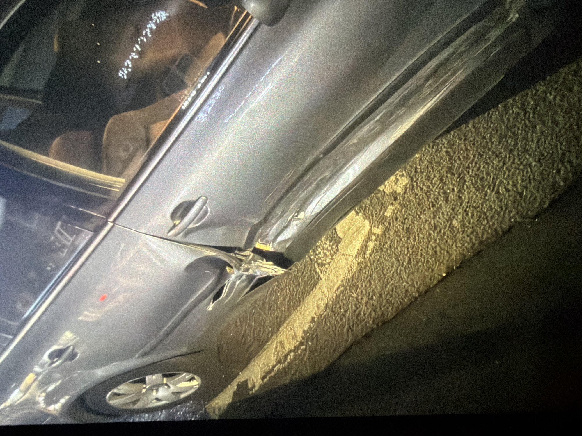 The image shows a close-up of the side of a silver car, with visible damage to the rear door and fender. The car is parked on a wet street, with reflections of lights on the surface of the road. A portion of a yellow painted line is visible on the road in the foreground. The scene appears to be a city street at night. The specific location in Sofia, Bulgaria, cannot be determined from the image alone.