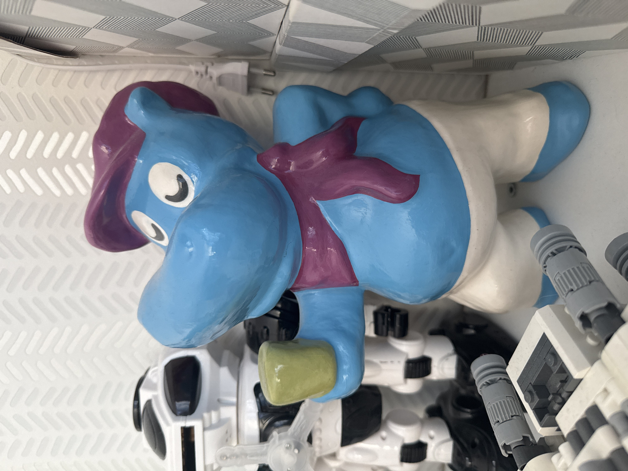 A blue, cartoonish hippo figurine with a purple hat and scarf, holding a small yellow cup, stands in the foreground. Behind it, partially visible, is a white and black robot toy. To the right of the hippo, a wall with a grey and white checkered pattern is visible, and further to the right, a white electrical plug is attached to the wall. In the bottom left corner, a close-up view of a white and grey Lego toy is present. The scene is indoors, with a white wall featuring a subtle chevron pattern in the background behind the robot. The overall setting appears to be a shelf or display area. The location is not specified within the image or its context.