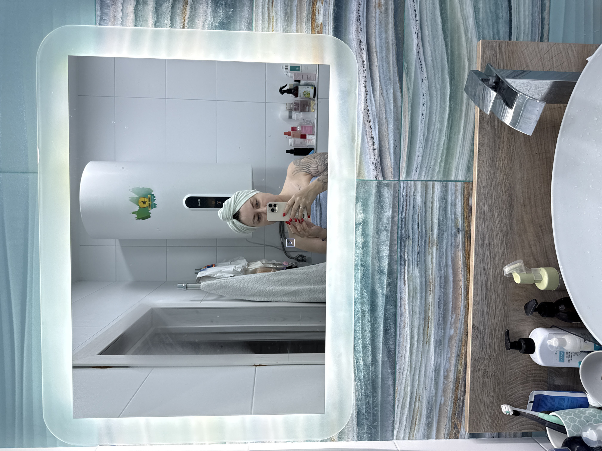 A person is taking a selfie in a bathroom mirror. The person is wearing a light green towel wrapped around their head and has tattoos on their arm. They are holding a white smartphone with a visible camera lens and are wearing red nail polish. The bathroom has white tiled walls and a white vertical water heater with a green and yellow sticker on it. In the reflection, there are several bottles and containers on a shelf above the water heater. The mirror itself has a rounded, illuminated frame. To the right of the mirror, there is a wooden countertop with a silver faucet and a white sink. Various toiletries, including pump bottles of soap and toothpaste, are on the countertop. The background features decorative wall tiles with a marbled pattern in shades of blue, green, and beige. The overall impression is a modern bathroom setting, possibly in Sochi, Russia. The time of day and weather are not discernible. The person appears to be casually posing for the photograph. Visible text includes "Haier" on the water heater and some text on a white pump bottle which appears to read "HAND".