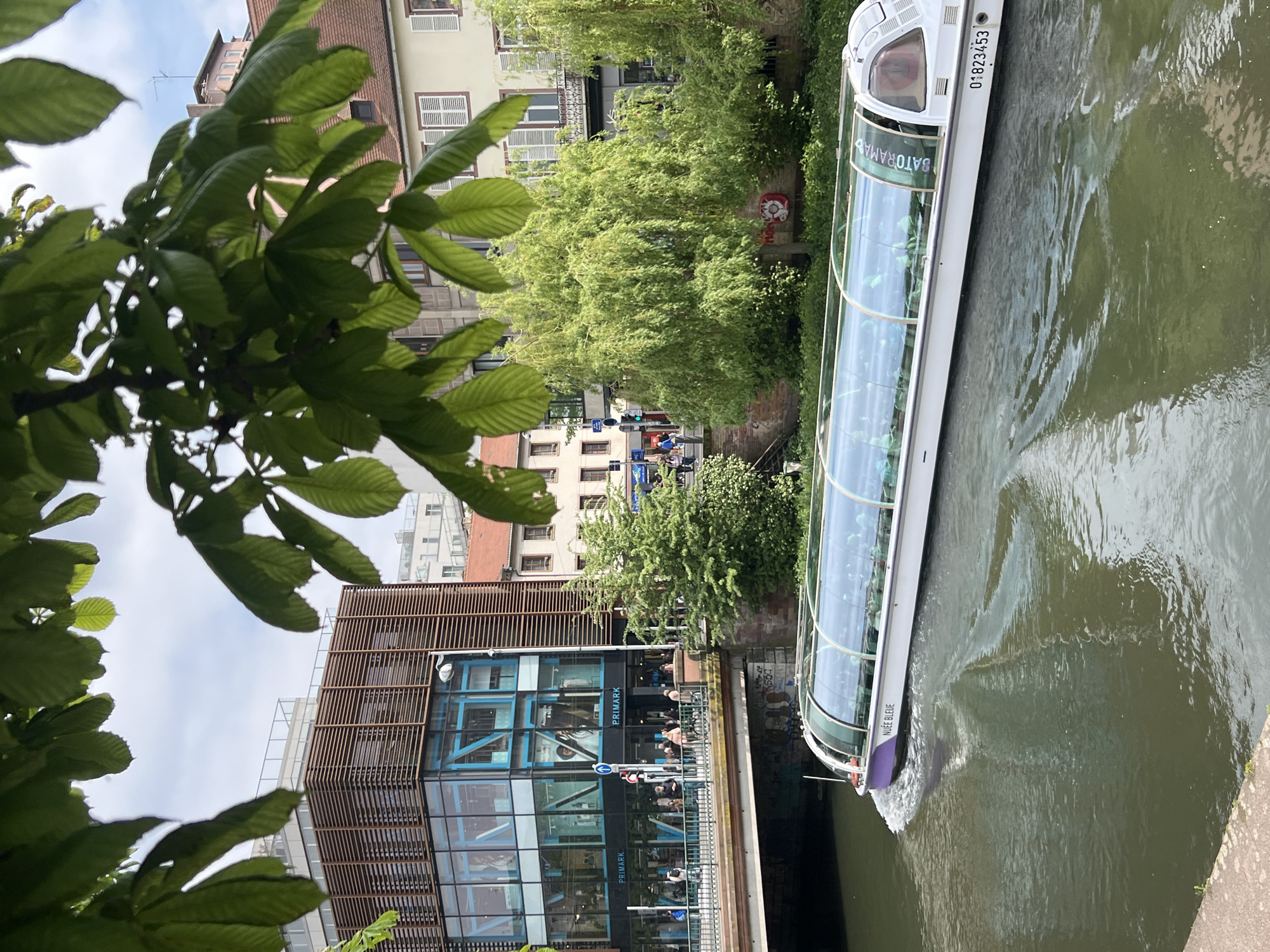 The image depicts a canal scene in Strasbourg, France. A Batorama tour boat navigates the canal. A Primark store is visible along the canal bank. People are present in front of the store. The environment includes buildings, trees, and foliage on the bank. The sky is partially visible through the tree leaves.