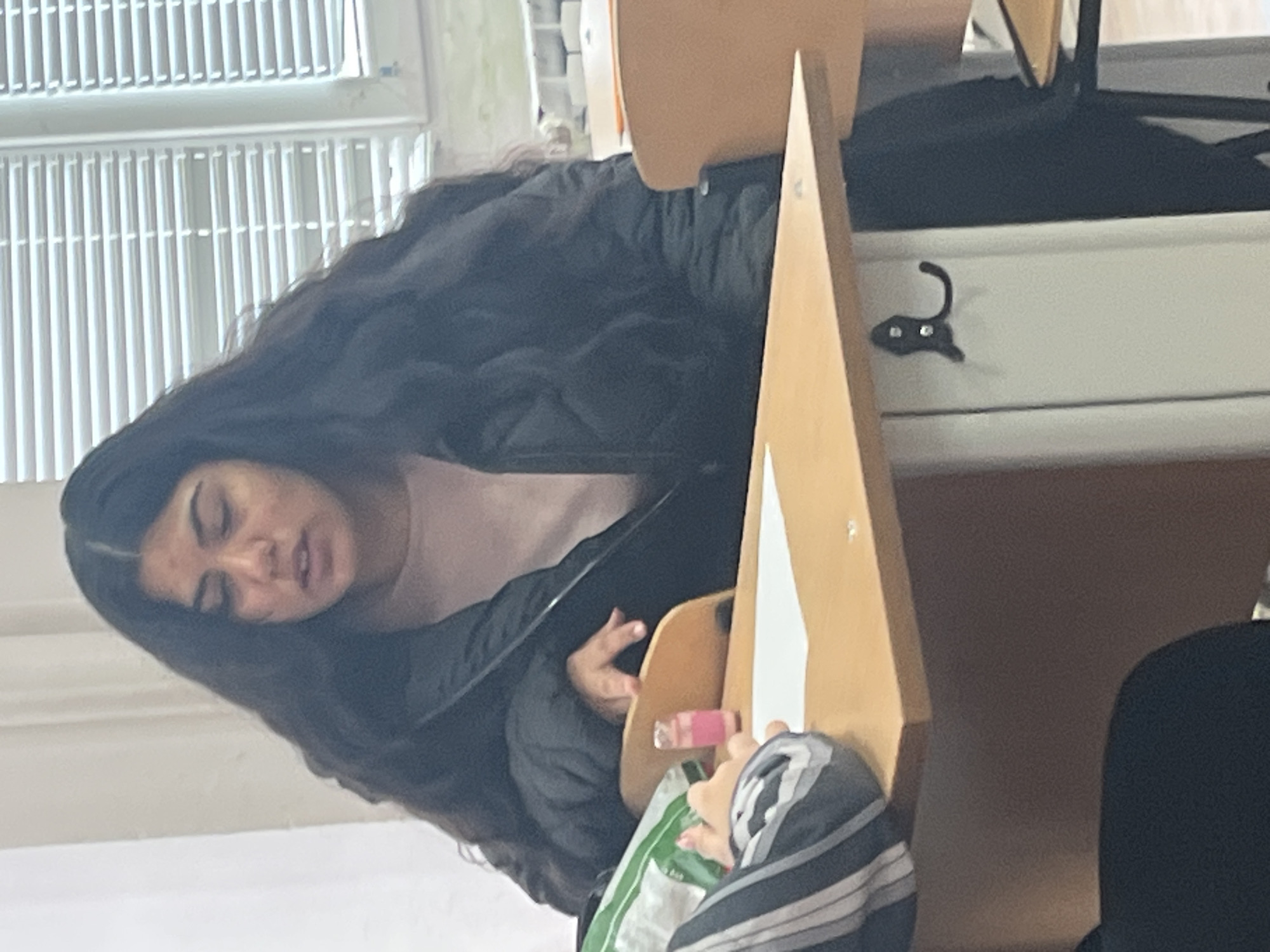 A young woman with long, dark hair is seated at a desk in what appears to be a classroom in Sofia, Bulgaria. She is wearing a light pink shirt underneath a dark jacket. The location is inferred from the style of the window blinds. The desk is a light wood color and has a folding flap attached. She is holding a small, pink item in her hand, possibly lip gloss. A striped scarf or bag is on the desk. Another person is partially visible to the right, wearing black clothing. A black hook is attached to the desk. The classroom is lit by natural light coming through the blinds.