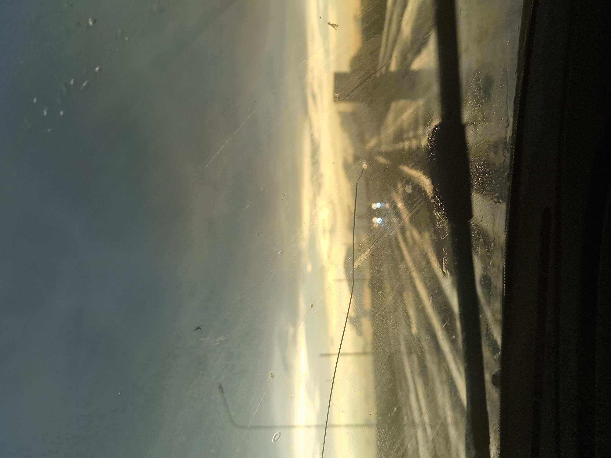 This image is a photograph taken from inside a vehicle, looking through a wet windshield at a highway scene at dusk. The sky is cloudy and appears to be a dark gray with streaks of light yellow and orange near the horizon, suggesting either a sunset or sunrise. The highway is wet, with reflections of lights and the sky on the road surface. There are what appear to be streetlights visible on the left side of the image. Two sets of headlights from approaching vehicles are visible in the distance on the highway. The windshield has numerous water droplets and some smudges, obscuring some of the details of the scene outside. The bottom of the image shows the dashboard and windshield wipers of the vehicle. The location is described as Unknown, Russia.