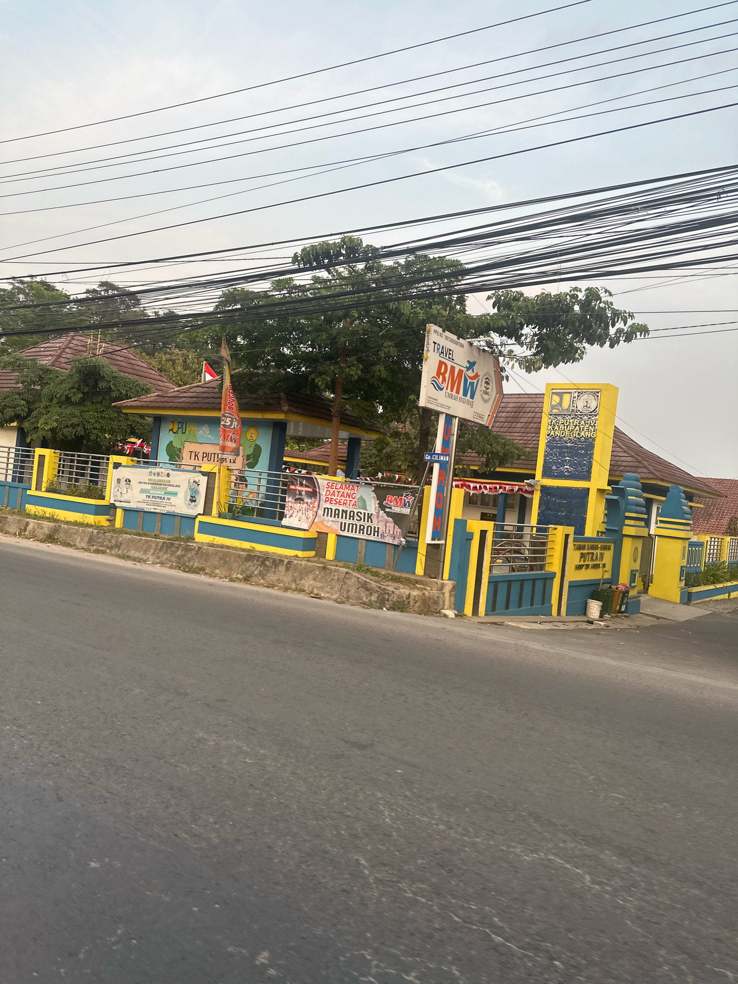 The image shows a street view in Saruni, Indonesia, dominated by a building with yellow and blue accents, a street sign, and overhead power lines. The building appears to be a school or community center, with "TK PUTRA IV" visible on a banner. Adjacent to it is a sign for "TRAVEL BMW" which offers "UMRAH AND HAJJ" services. Another prominent sign with yellow and blue colors indicates "KEMENAG KABUPATEN PANDEGLANG," suggesting it's related to the Ministry of Religious Affairs in Pandeglang Regency. The road in the foreground is asphalt, and trees and vegetation surround the building. The sky is overcast.