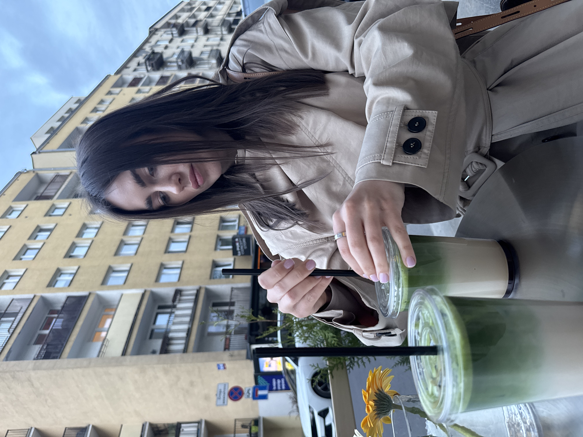 A medium shot depicts a young woman seated at an outdoor table in Warsaw, Poland. She is wearing a beige trench coat and has long, dark hair. She is stirring a beverage with a black straw in a clear plastic cup. Next to it, another similar drink. A small vase containing a yellow flower is also on the table.
Behind her, a multi-story building can be seen, characterized by numerous windows. Street signs and a white car are visible in the background. The scene appears to be a casual urban setting, with the woman enjoying a drink at an outdoor cafe or restaurant.