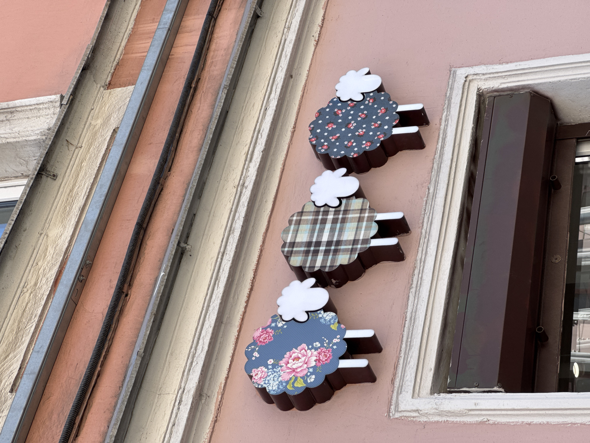 This image provides a low-angle, close-up view of a building facade in Saint Petersburg, Russia. The wall is painted a light salmon pink. At the bottom of the frame, a white, decorative architectural molding surrounds the top portion of a window or door opening, which is fitted with a dark brown rolling shutter.
Above the window frame, three stylized sheep-shaped signs are mounted horizontally on the pink wall. Each sign consists of a dark brown, undulating three-dimensional body, a flat patterned panel representing the fleece, a white cloud-shaped head, and two white rectangular legs.
From left to right:
1. The first sheep's fleece is dark blue with a floral pattern featuring large pink peonies, smaller blue flowers, and green leaves.
2. The second sheep's fleece displays a brown, beige, and light blue plaid pattern.
3. The third sheep's fleece is dark blue or gray with a repeating pattern of small red flowers and white dots.
Above the sheep signs, a corrugated black electrical conduit runs horizontally across the pink wall. Further up, a metallic gray channel is visible, topped by a weathered beige architectural band. The scene is illuminated by bright, even natural light, indicating daytime. The building facade exhibits some minor signs of wear and slight discoloration.