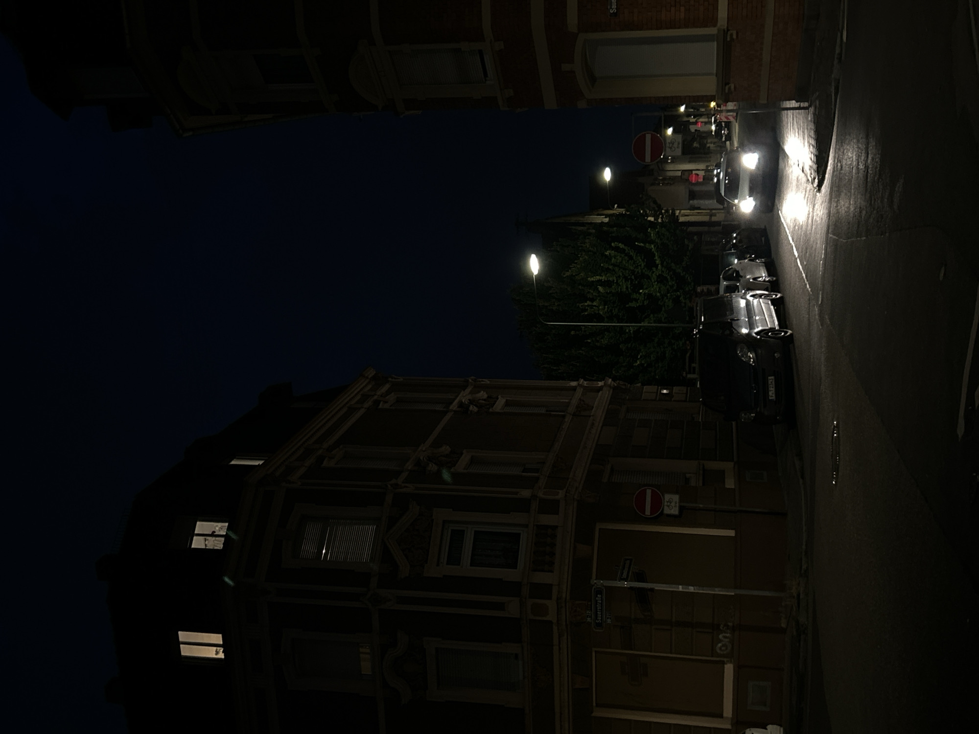 The image depicts a nighttime street scene in Frankfurt, Germany. A dark blue sky is visible above dark buildings lining a street. On the left, a multi-story building with illuminated windows and ornate architectural details stands. Across the street, a red brick building is visible on the right.
Several vehicles are present. In the foreground, a white car with its headlights on is driving down the street, casting bright reflections on the wet asphalt. To the left of this car, other vehicles, including a dark-colored van and at least two other cars, are parked or moving.
Streetlights illuminate parts of the scene, and a traffic sign with a red circle and a bicycle symbol is mounted on a pole near the intersection. The overall atmosphere is one of a quiet urban evening.