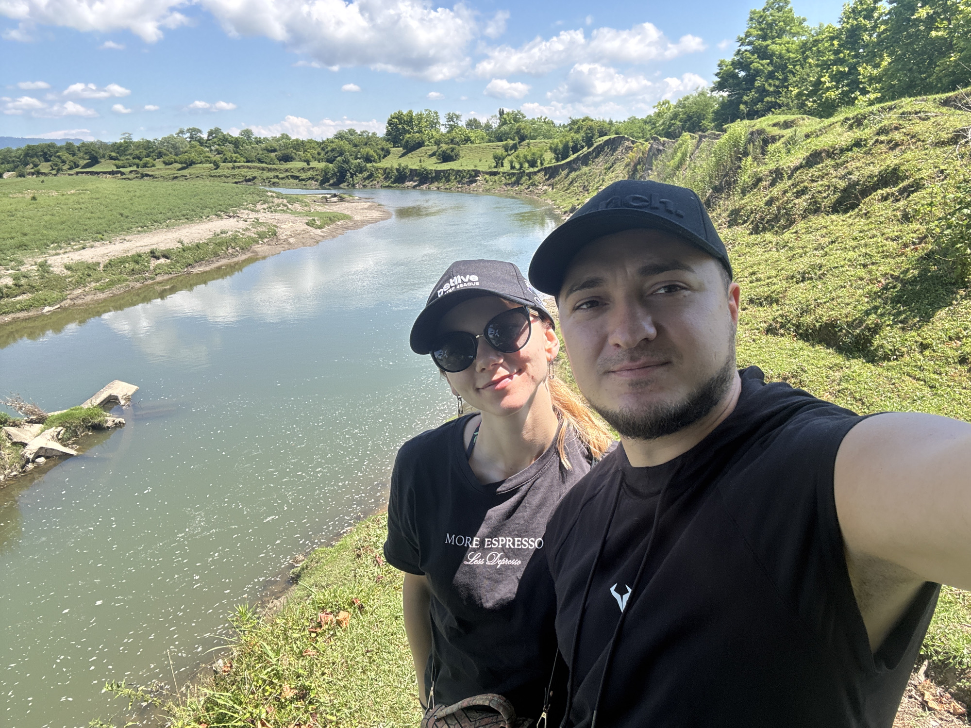 In the city of Darcheli, Georgia, a man and a woman are captured in a selfie outdoors. The couple stands on the grassy bank of a wide, calm river, with lush green hills and trees forming the backdrop. The woman, wearing sunglasses and a black t-shirt with white lettering that reads "MORE ESPRESSO Less Depresso", is positioned to the left of the frame. She has blonde hair tied back and is smiling. The man, wearing a black baseball cap and a black t-shirt with a white logo, is on the right, looking towards the camera with a slight smile. The river, with its greenish-blue water, reflects the sky and surrounding vegetation. In the distance, a small, dilapidated wooden structure is visible on the riverbank. The sky is a bright blue, dotted with fluffy white clouds, indicating a pleasant, sunny day. The overall scene conveys a casual and relaxed atmosphere.