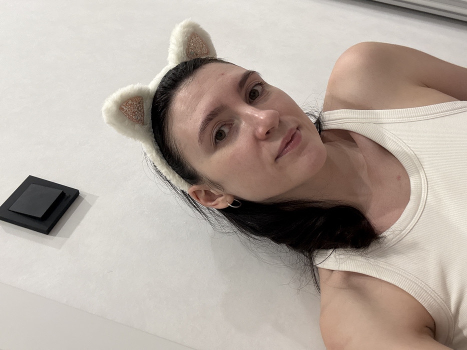 A woman lies on a white surface wearing a white ribbed tank top and a cat ear headband. Her dark hair is partially visible. She looks toward the camera with a subtle smile. To the left, near her head, is a square black object on the white surface. The lighting appears soft, with minimal shadows. The woman's head is angled slightly.