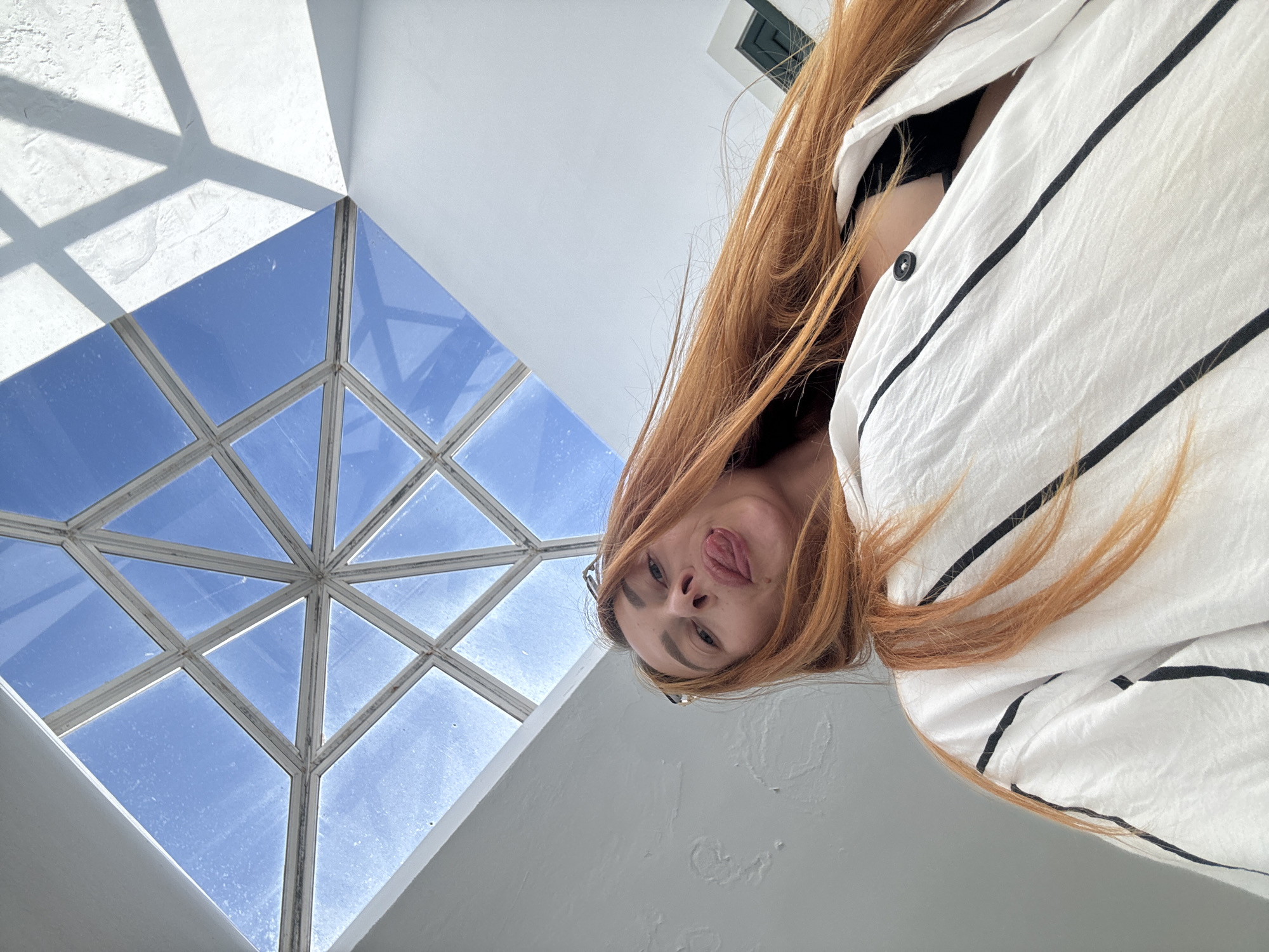 In a wide-angle, low-angle selfie taken from within a building in Budva, Montenegro, a woman with long, vibrant auburn hair sticks her tongue out playfully towards the camera. She is wearing a white, loose-fitting shirt with vertical black pinstripes and a black bra visible underneath. Her hair is slightly windblown, framing her face. Above her, a striking geometric skylight with a diamond pattern of white frames allows bright blue sky to filter into the space. The walls of the interior are painted white, with one wall showing a rough texture. Shadows from the skylight's frame are cast across the textured wall, creating a pattern of light and shadow.