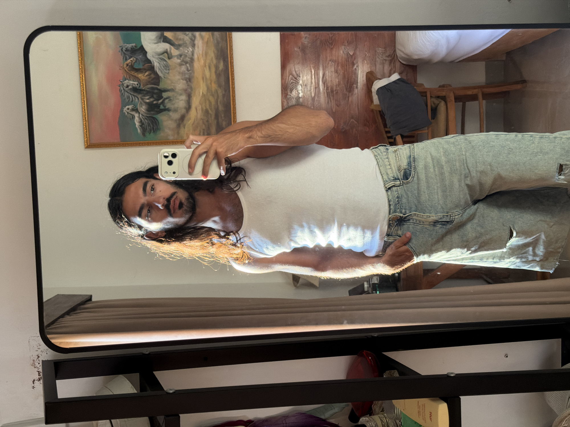 A full-length mirror selfie captures a person with long dark hair, a beard, and a mustache, standing indoors in North Kuta, Indonesia. The person wears a white ribbed tank top and light blue distressed denim shorts with a rip on the right leg. They hold a light-colored smartphone with a red-painted fingernail visible, taking the photo directly in front of the mirror.
Strong natural sunlight illuminates the scene, creating bright highlights on the person's hair and clothing, suggesting it is daytime. The person maintains a neutral expression while looking at the phone.
Reflected in the mirror, behind the person's upper body, is a framed painting depicting several wild horses in motion against a warm-toned, possibly sunset-colored, sky. To the right of the person's reflection, elements of the room are visible, including wooden wall paneling, a wooden chair with dark clothing draped over it, and a white bed with white bedding.
Below the mirror, there is a dark-framed shelving unit. Various items are partially visible on these shelves, including a red object, a white roll, and a yellow block object with the text "Nur" and "Makeup" printed on it. The white wall surrounding the mirror shows some minor peeling paint in the bottom left corner. The overall setting appears to be a casual indoor space, likely a bedroom.