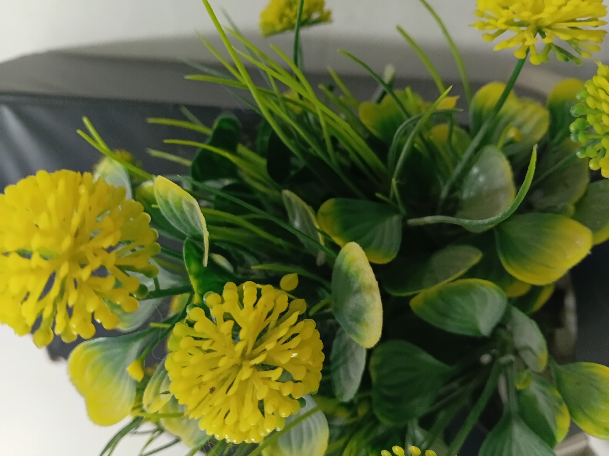 A close-up shot showcases a bouquet of artificial yellow flowers with green leaves. The flowers are vibrant yellow, with many small, petal-like structures clustered together to form a rounded bloom. The leaves are a mix of green and yellow, with some appearing more faded than others. The background is a blur of dark gray and white, suggesting an indoor setting, possibly a home or office in Wibawamulya, Indonesia. The lighting is soft, highlighting the textures and colors of the artificial flora. There are no people or animals visible in the frame.