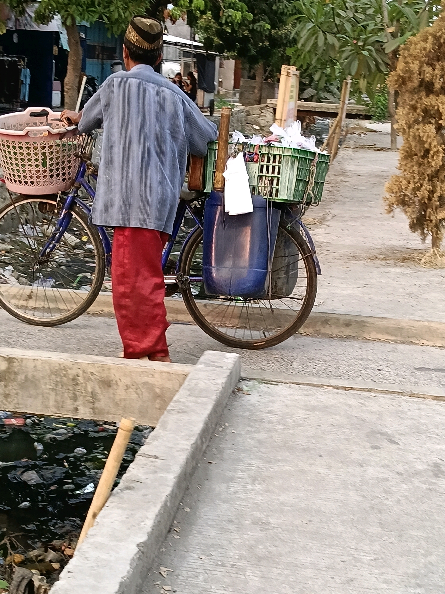 A person wearing a striped blue shirt and red pants stands with their back to the camera, holding onto the handlebars of a blue bicycle. The bicycle has a pink basket attached to the front and a green plastic crate secured to the back. Inside the crate are white plastic bags. A large blue plastic container is also attached to the rear of the bicycle. The person appears to be in an urban or semi-urban environment. In the foreground, a concrete structure borders a drainage ditch filled with dark water and debris, including plastic waste and leaves. The ground surface is a mix of paved concrete and dirt. In the background, there are buildings and trees. The scene is consistent with the city of Belimbing, Indonesia, a location known for its urban development and environmental challenges related to waste management.