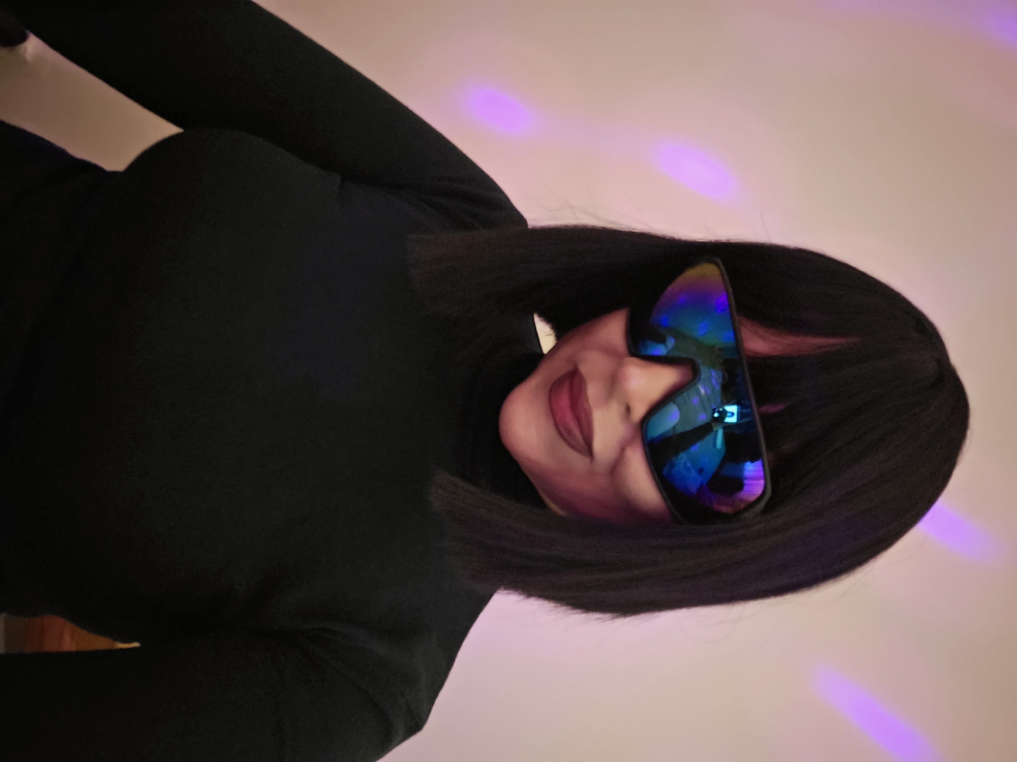 A close-up portrait features a person, likely female, with short, straight black hair styled in a bob cut, wearing a black turtleneck top. The individual's head is tilted slightly, and they appear to be looking towards the viewer. Their eyes are concealed by large, wide-framed, futuristic-looking sunglasses with highly reflective lenses that shimmer with vibrant blue and purple hues. A subtle smile is visible on their lips, which are painted a dark pink or plum color. The background consists of a light-colored surface, possibly a wall, illuminated by distinct splotches and streaks of purple and pink light, suggesting an indoor setting with atmospheric or club-like lighting, indicative of evening or night. The hair has a uniform, blunt appearance, resembling a wig. No text is visible in the image. The overall impression is one of modern, somewhat mysterious style.