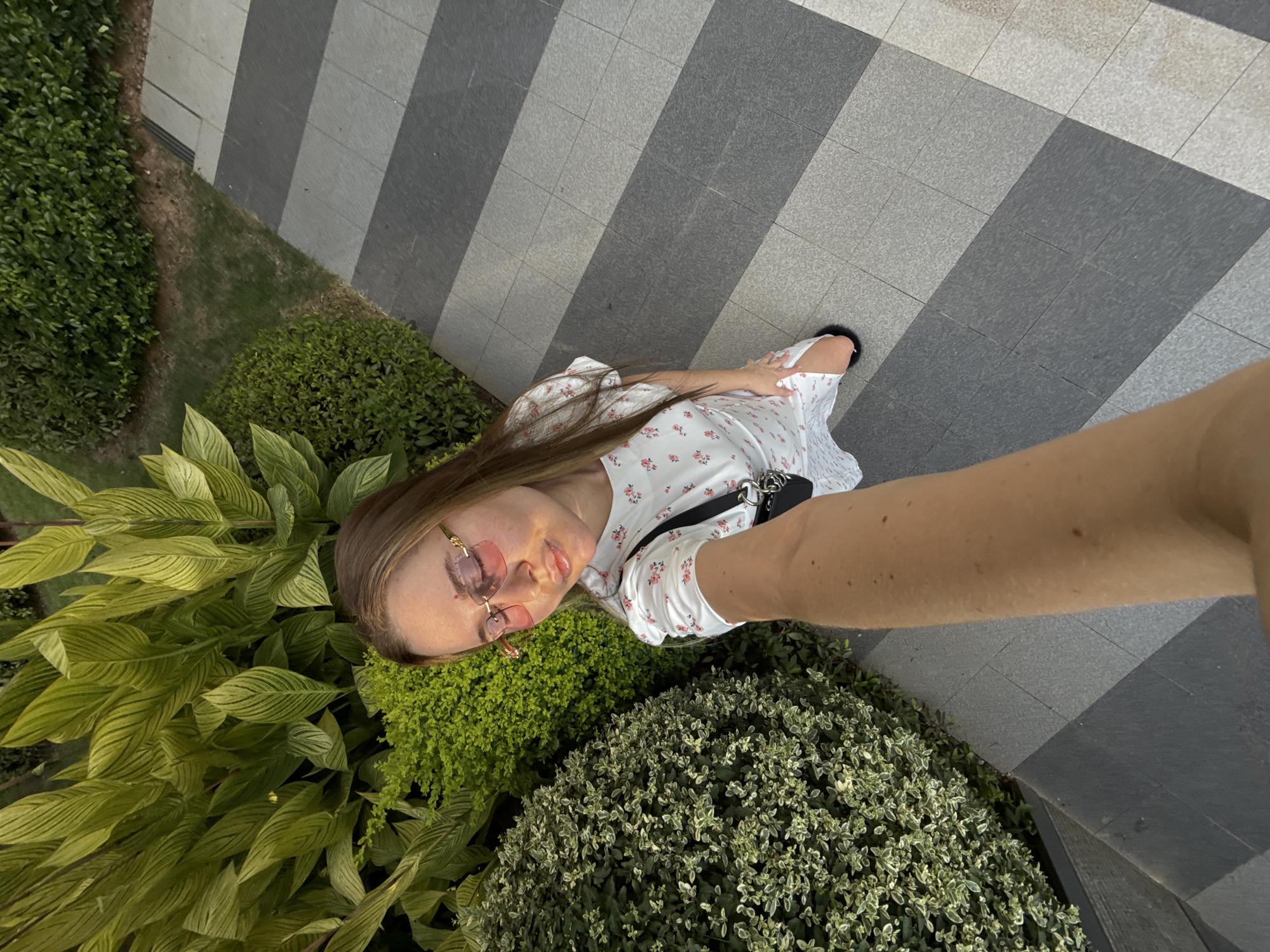 A woman is taking a selfie outdoors, standing on a paved walkway with a pattern of gray and lighter gray tiles arranged diagonally. To her left is a lush green garden with various types of plants, including large, striped leaves and smaller, dense bushes. She is wearing a white dress with a small floral pattern, pink-tinted rectangular glasses, and a black crossbody bag with a silver chain detail. Her arm is extended, holding the camera, and her body is angled slightly away from the camera as she looks down. The lighting suggests it is daytime, with sunlight casting shadows. The environment appears to be a well-maintained outdoor space, possibly a park or a garden area within the city of Yiwu, China.