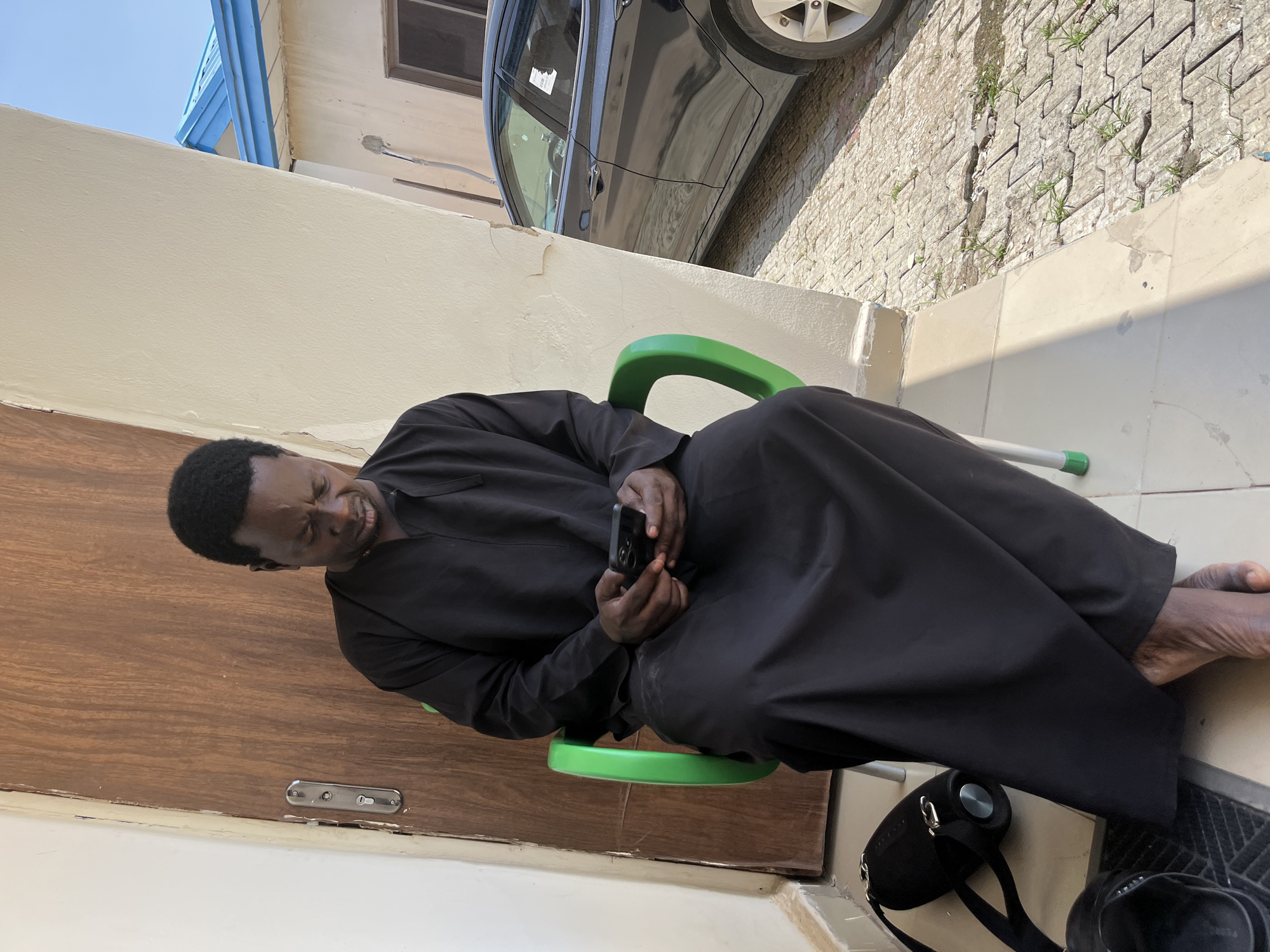 A Black male in his 50s, wearing a black religious robe, is seated on a green plastic chair outdoors, looking down at a smartphone in his hands. He is positioned in front of a wooden door with a silver lock mechanism. To his right, a light-colored wall curves away, revealing a portion of a black car parked on a paved surface. The environment suggests a residential or public area in Abuja, Nigeria, with the sun casting shadows on the ground. A black bag and a pair of dark sandals are visible on the ground near the man's feet. The scene is captured in daylight.