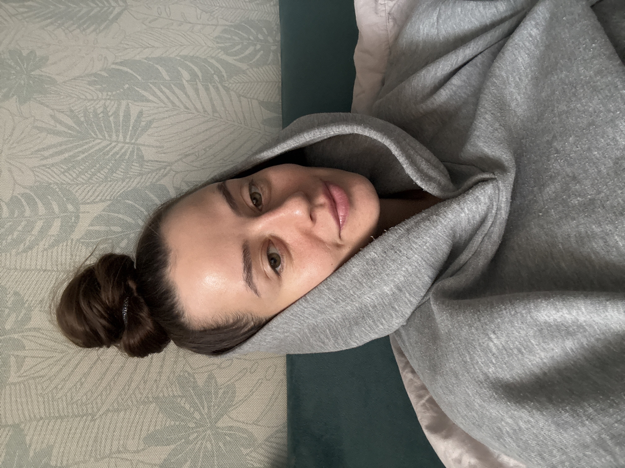A young woman with light skin, brown hair in a bun, and green eyes is lying down, wearing a gray hooded sweatshirt. The hood is pulled up around her face. She is looking directly at the camera with a slight smile. The background includes a patterned wallpaper in muted green and white tones, a dark green headboard, and a glimpse of a light pink or beige bedding. The location is not identifiable from the image.