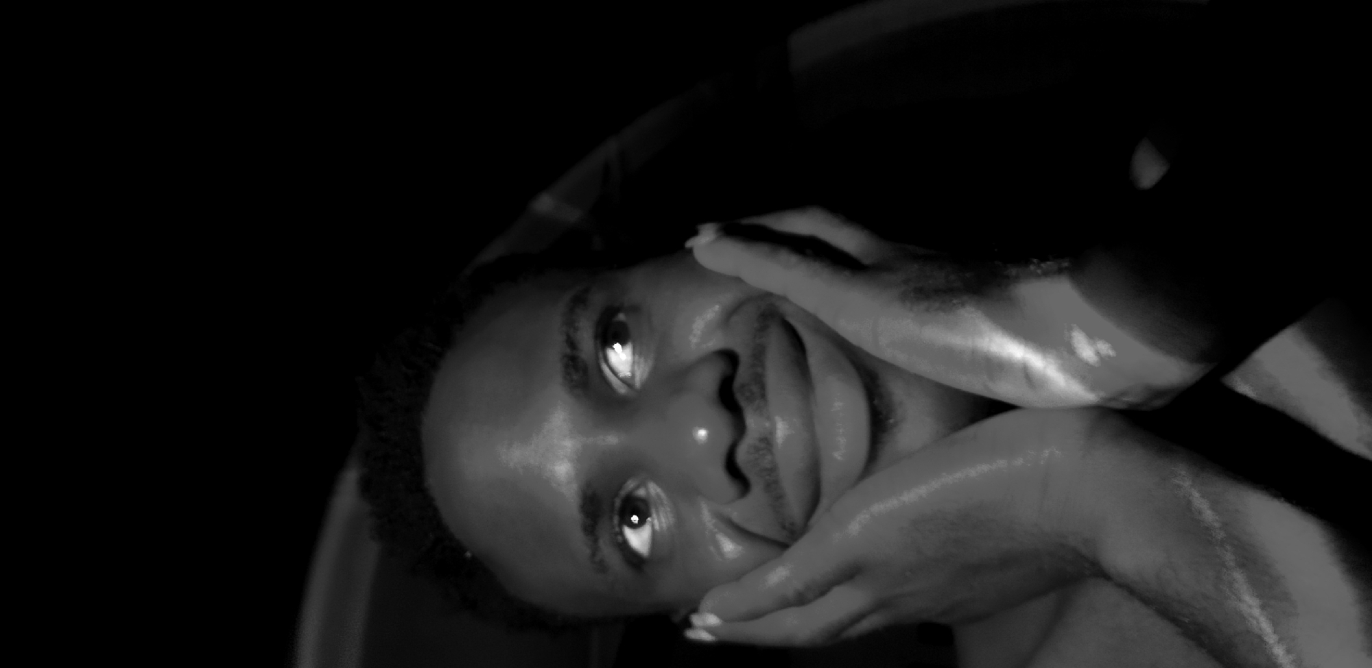 The image shows a close-up, black and white shot of a young Black man's face, with his hands placed on his cheeks. He has short, curly hair and is looking upwards and to his right with his eyes wide open. His lips are parted in a slight smile, and there's a mustache above his upper lip. The lighting is dramatic, with strong highlights on his face and hands, and the background is almost entirely black, suggesting a dark environment. The man appears to be indoors, possibly in a car or a room with minimal lighting. The scene is posed and appears to be a self-portrait or a casual photograph. There are no discernible elements in the image that would indicate a specific location in Narayi, Nigeria, or any urban setting.