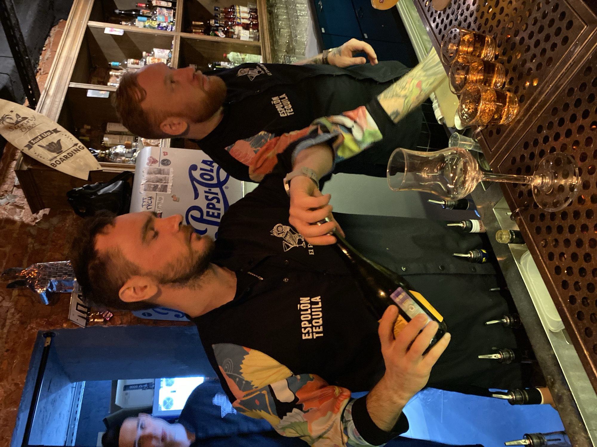 Two men are working behind a bar, possibly in a bar or restaurant in Izhevsk, Russia. The man in the foreground holds a bottle of Espolòn Tequila. A series of shot glasses and martini glasses line the bar surface. There is a Pepsi-Cola cooler. A surfboard-shaped sign with the words "Sunny Day Surf Up BOARDING" is visible. The two men wear black shirts with a logo and the text "ESPOLON TEQUILA." The background includes shelves filled with liquor bottles. The image may be rotated sideways.