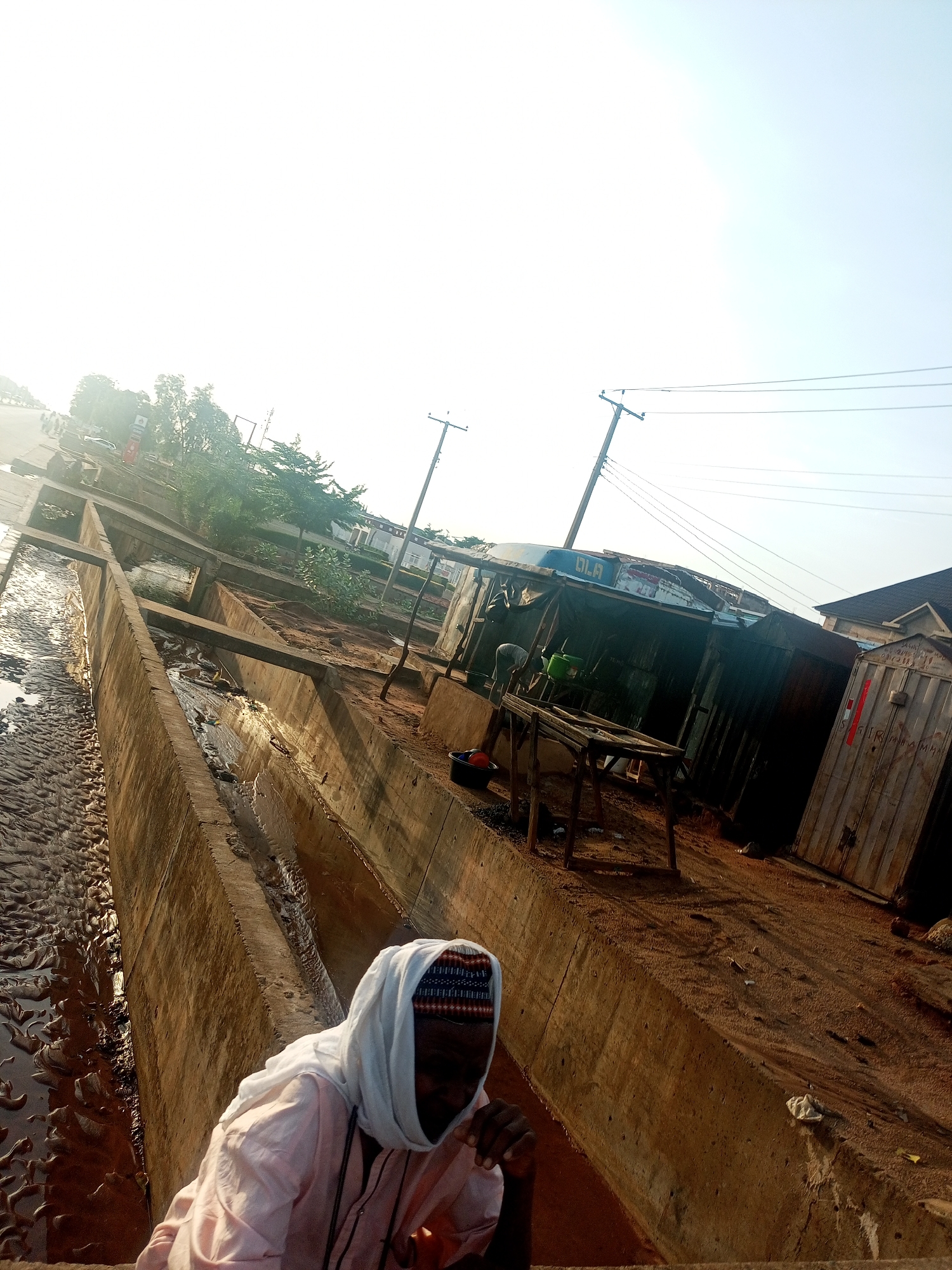 A heavily tilted outdoor image depicts a sunlit, somewhat rugged urban or semi-urban environment, likely taken during early morning or late afternoon due to the strong, low-angle light. The sky is very bright, almost blown out.
In the foreground, a person is partially visible, seen from an elevated perspective looking down. They wear a light-colored head covering or scarf over a cap with a dark, patterned band. Their face is largely obscured by shadow. They are dressed in a light-colored, long-sleeved shirt, possibly pink or peach. Their right hand is near their chest.
The most prominent feature in the midground is a wide, multi-tiered concrete drainage ditch or canal. The leftmost section of the ditch contains dark, muddy, and textured ground, possibly where water has receded, with reflections of the bright sky on remaining puddles. The inner sections of the concrete ditch contain murky, brownish water and some debris.
To the right of the concrete ditch, there's a dirt path or ground area leading to several rudimentary structures. These appear to be makeshift shelters or stalls constructed from corrugated metal sheets and other materials. One of these structures has the word "OLA" written in blue capital letters on its side. Another structure, resembling a container, has "IRFAMMAM" partially visible. Near these structures, a simple wooden frame or table stands, and a person in light clothing appears to be engaged in an activity, with a green container nearby.
In the background, utility poles with overhead power lines stretch across the scene. Further back, on what appears to be a paved road, indistinct vehicles and some vegetation are visible. The overall scene suggests a functional, possibly informal, settlement area with a focus on practical infrastructure like the drainage system and power lines.