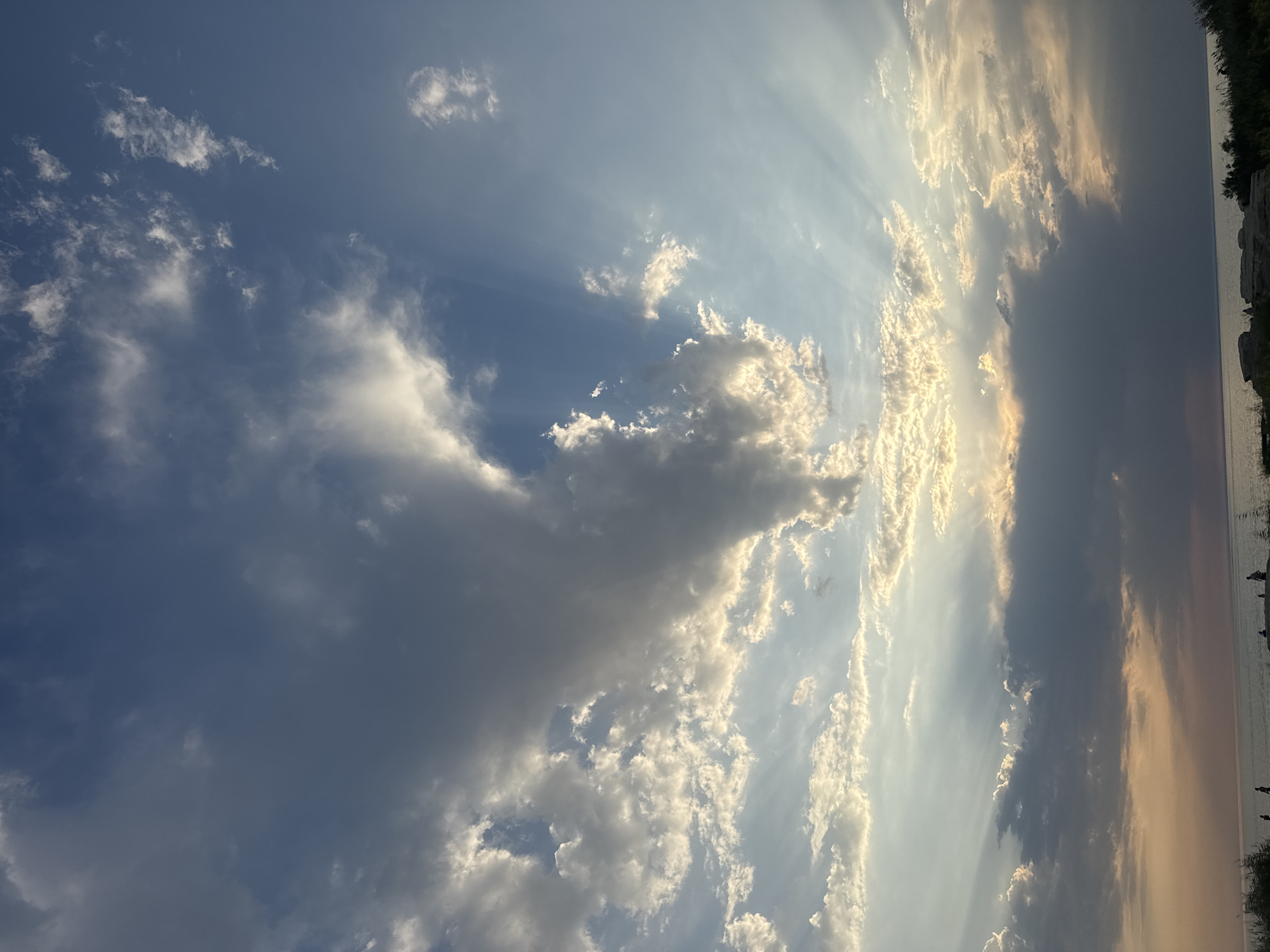 The image depicts a dramatic sky with crepuscular rays shining through scattered clouds. The sky transitions from a deep blue at the top to lighter shades near the horizon, where a body of water, likely the Caspian Sea, is visible. Along the shoreline, silhouetted figures can be seen, suggesting a public space such as a beach or promenade. The overall atmosphere is serene, with the setting sun casting a warm glow. The location is identified as Aktau, Kazakhstan.