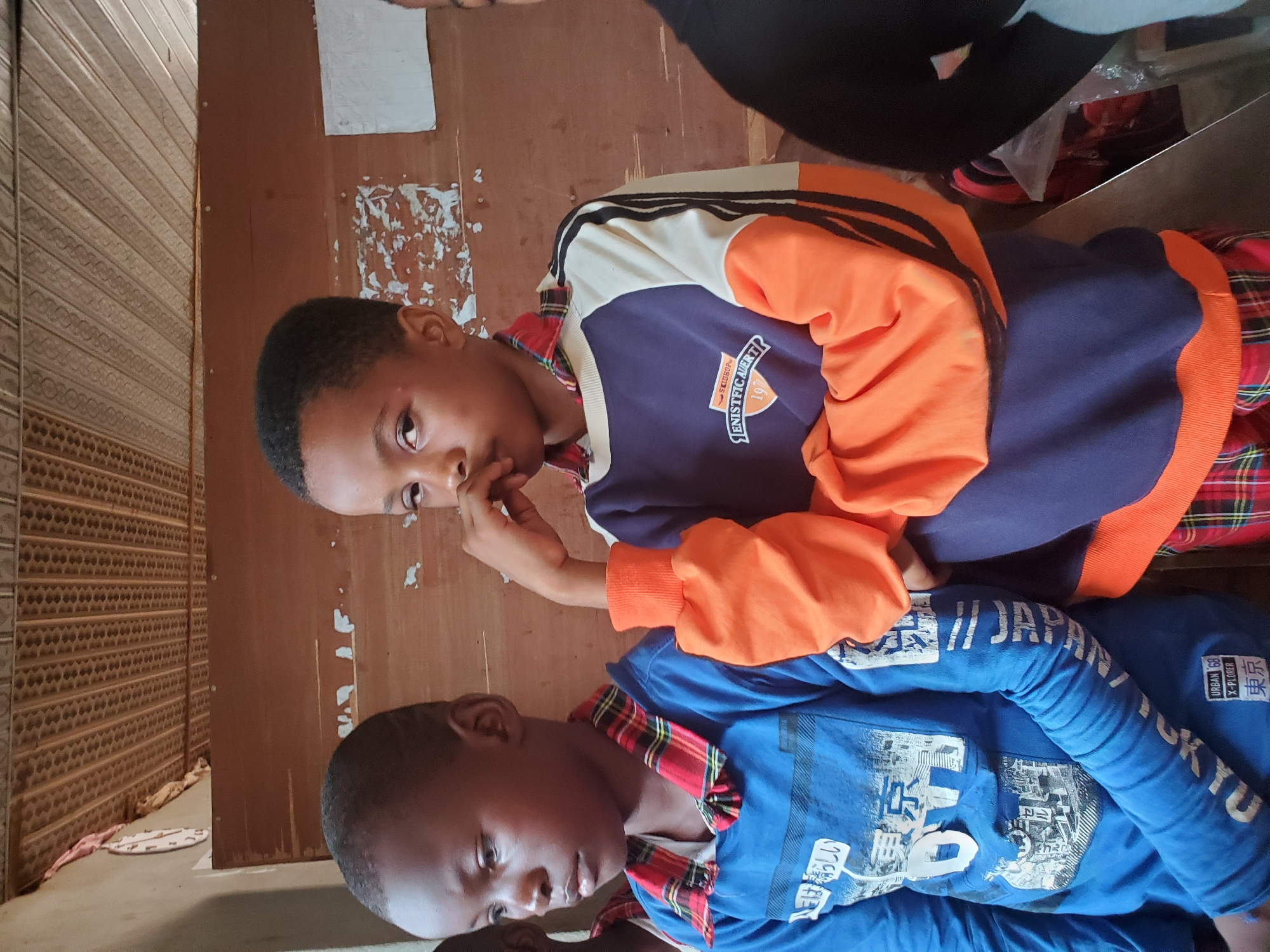 This image, captured in Ukwu-Abwa, Nigeria, displays two young boys in an indoor setting, with the photograph oriented sideways.
On the left side of the frame, a boy wears a blue long-sleeved shirt featuring white Japanese text and graphics. Visible text elements on his shirt include "には晴らしい", "東京" (Tokyo), "TOKYO", "URBAN 68", and "X-PLORER". He also has a red and black plaid collar. His gaze is directed slightly downwards, conveying a calm expression.
To his right, another boy is clad in a distinctive long-sleeved shirt or jacket, characterized by a color-block design in navy blue, bright orange, and white, with black stripes adorning the shoulders. This garment also features a red and black plaid collar. Text visible on his clothing includes "SKIDHOP™", "ENISTFIC AVERT", and the number "197". He looks directly towards the camera, with one hand raised towards his mouth, suggesting a thoughtful or perhaps shy demeanor.
The background primarily consists of a plain brown wooden wall, exhibiting signs of wear such as peeling paint or residue. To the far left, a different textured, patterned wall or door panel is visible. A person's head and shoulder are partially visible in the top right corner of the frame, indicating other individuals are present. The overall setting appears to be a casual, well-worn indoor space, and the image captures a candid moment between the boys.