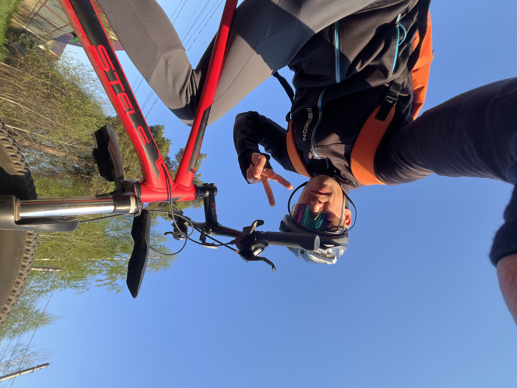 A low-angle shot shows a cyclist astride a red and black STELS bicycle, oriented upside-down in the frame. The cyclist, wearing a black and orange jacket, gray pants, and a helmet with a visor, makes a peace sign with their left hand. The background consists of a clear blue sky above a blurred landscape of green trees and bushes. The location is speculated to be Chaikovsky, Russia. The bicycle features a front suspension fork with a black mudguard. The cyclist's jacket has the brand name "NORDSKI" visible on the sleeve.