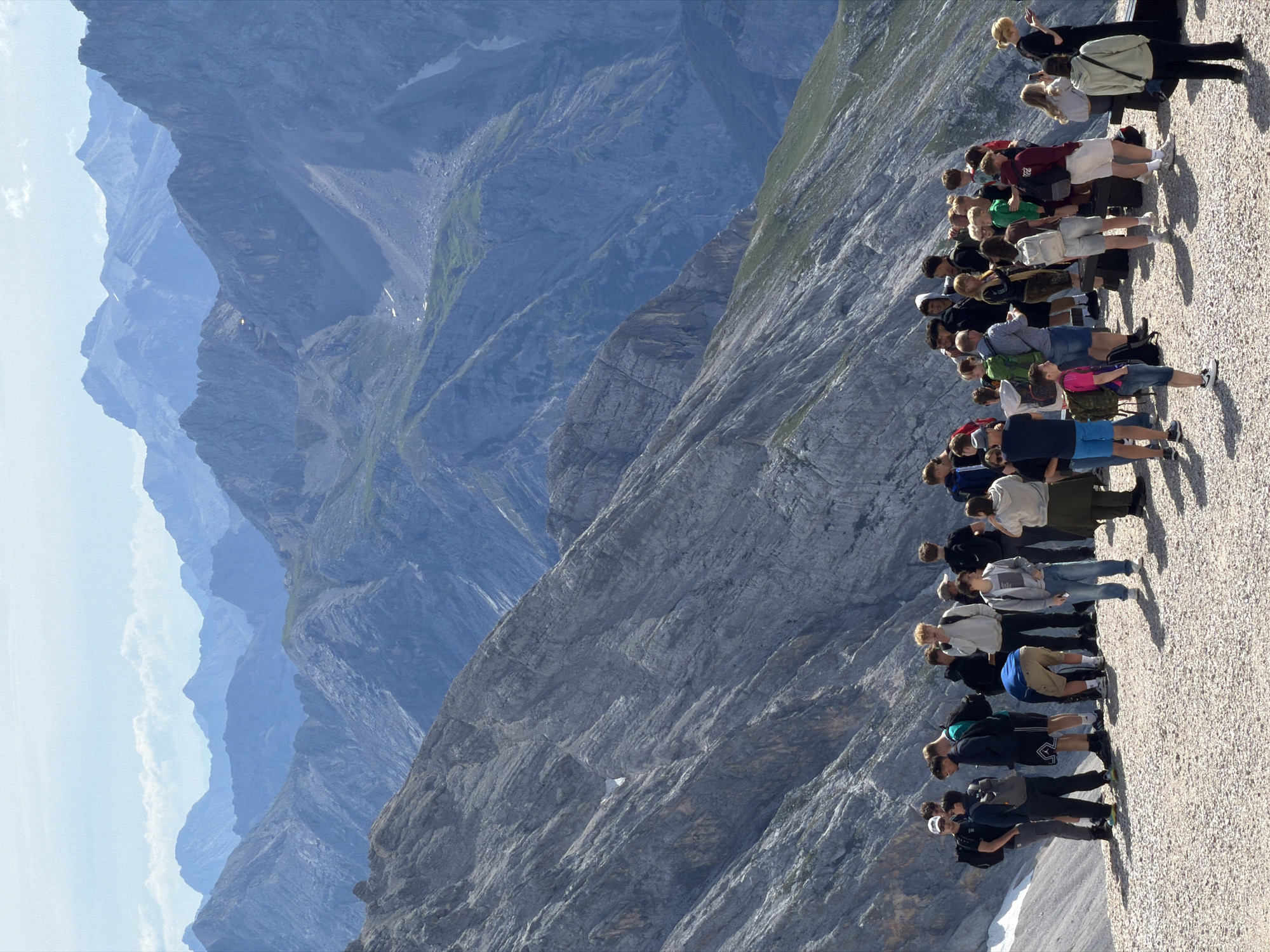 The image depicts a group of approximately 30 individuals on a rocky, gravel-covered vantage point, likely a mountain peak or observation deck, overlooking a vast, layered mountain range in the Bavarian Alps. The mountains in the background are predominantly shades of blue and grey, with visible ridges, valleys, and some patches of green vegetation and snow. The sky is a pale blue with scattered white clouds.
The people are dressed in casual, outdoor attire, suggesting a tourist or hiking group. They are standing and interacting, with some looking out at the scenery, others conversing, and a few engaged in taking photos or looking at their phones. The individuals vary in age, with a majority appearing to be young adults and teenagers.
Several people are wearing backpacks, and one individual is holding what appears to be a map or brochure. The ground they are standing on is uneven and covered with small stones and pebbles. Shadows are cast on the ground, indicating a sunny day.
While the exact location within Garmisch-Partenkirchen is not explicitly shown, the majestic mountain panorama strongly suggests a location with a view of the Wetterstein Mountains, such as the Zugspitze or a nearby viewpoint accessible from the town. The presence of tourists in this scenic alpine setting is characteristic of the popular tourist destination.