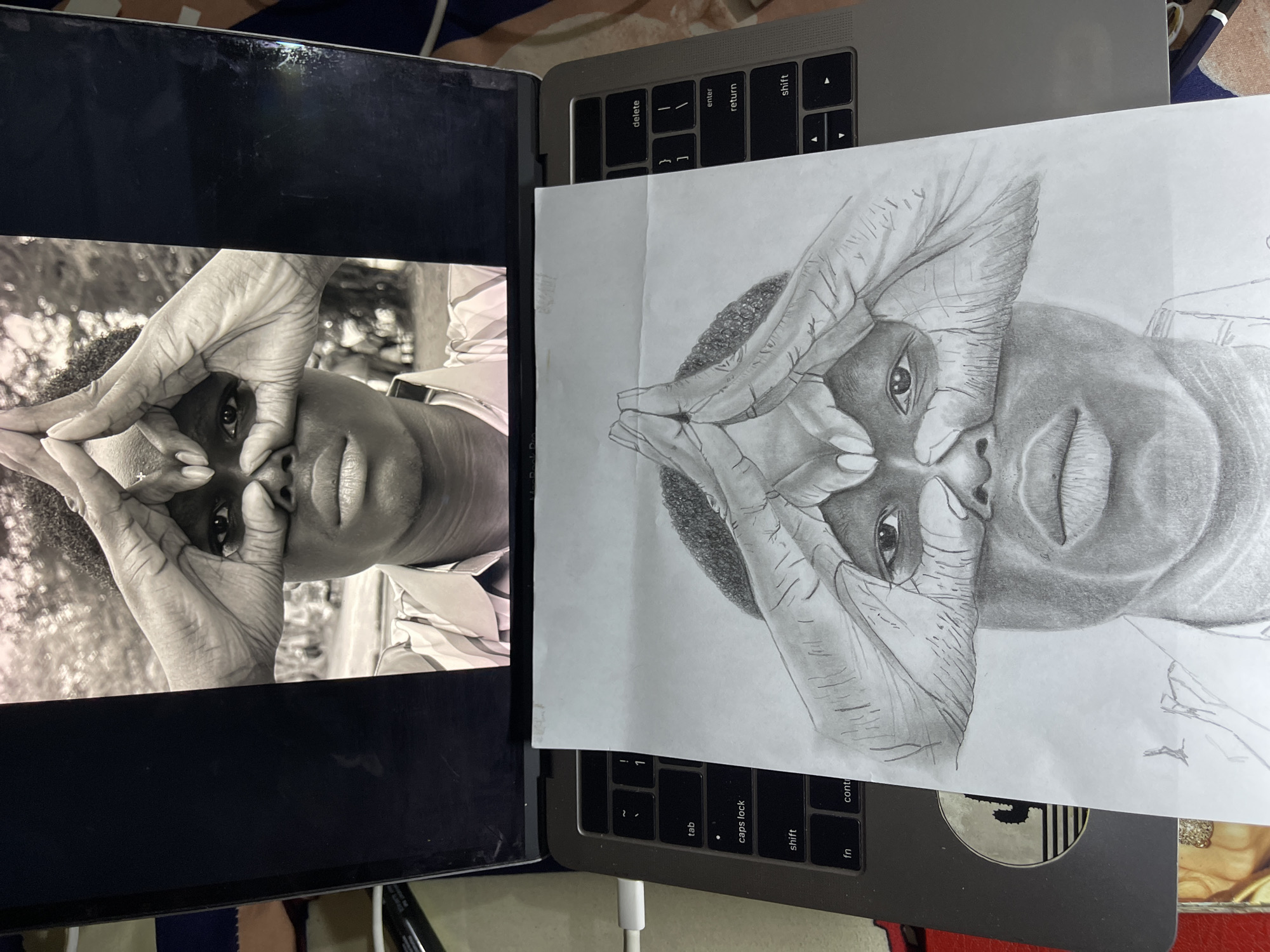 A drawing on white paper is partially covering a laptop keyboard. The drawing is a portrait of a person shown in a similar pose in a black and white photograph. The photograph is on a black frame. The image is not from Jos, Nigeria.