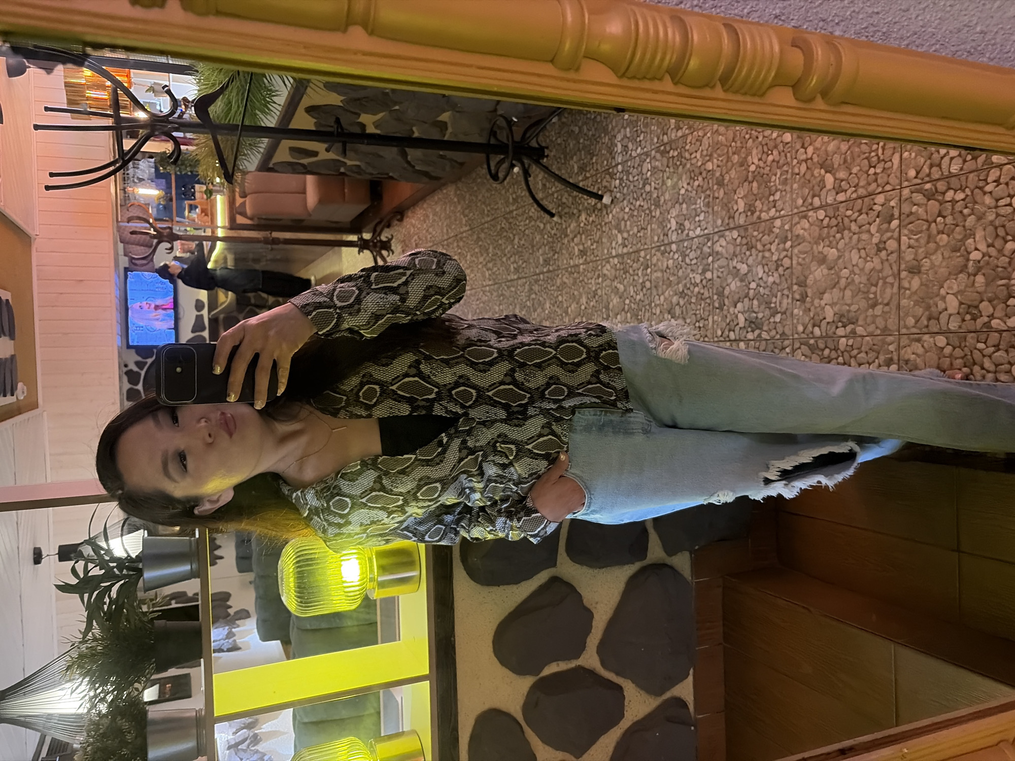 The image is a vertical, slightly distorted selfie taken in a room with rustic decor. A young woman with long dark hair is in the foreground, holding a smartphone to take the picture. She is wearing a snakeskin-patterned long-sleeved shirt and ripped light-wash jeans. Her expression is neutral.
Behind her, the reflection in a large mirror reveals a seating area with plush couches and a television screen displaying an image of a pink-haired person. The walls are a mix of light-colored wood paneling and textured stone tiles. Green potted plants are visible on shelves, and yellow lamps provide warm lighting, suggesting the scene takes place indoors. The overall ambiance is casual and relaxed. No visible text is present in the image.