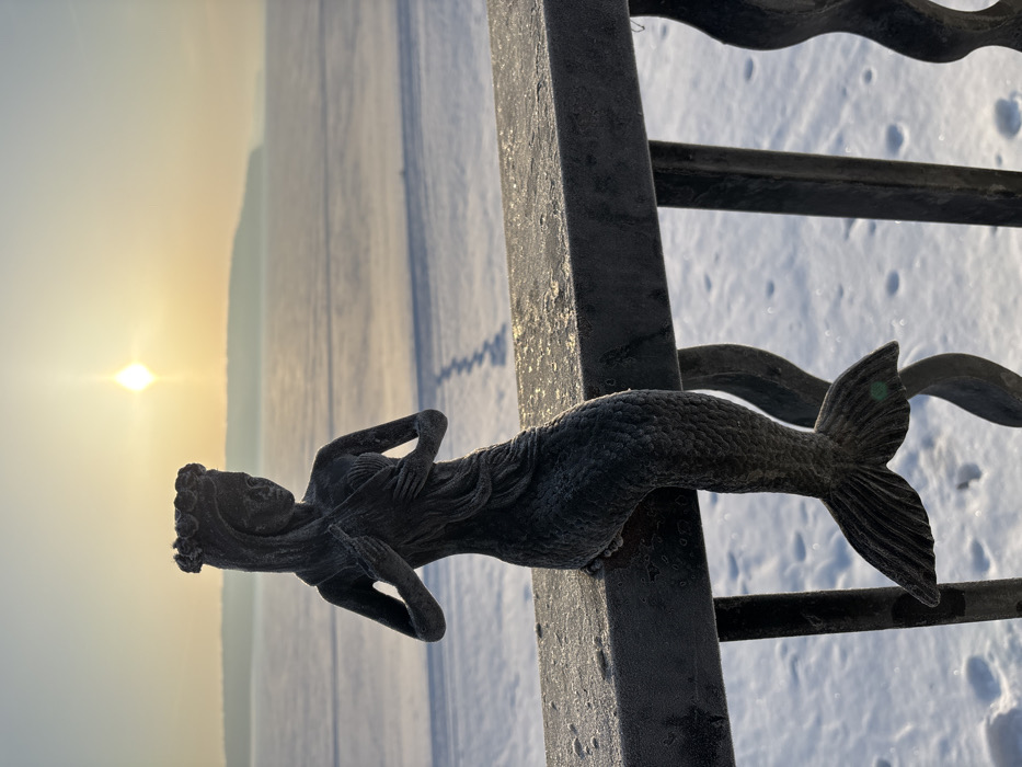 A weathered, dark metal statue of a mermaid is perched on a frost-covered black metal railing, facing slightly left. The mermaid wears a crown of what appears to be leaves or flowers, has long hair, and holds her hands near her chest, partially covering her torso. Her scaled tail extends downwards, ending in a prominent fluke. The railing, exhibiting visible ice or frost accumulation, features decorative vertical scrolls beneath its horizontal bar.
The scene is set outdoors in a winter environment during either sunrise or sunset, indicated by a low, bright sun with a visible flare in the upper center-right of the frame. Beyond the railing, a blanket of undisturbed snow covers the ground, with a distinct set of footprints trailing diagonally from the middle-ground towards the distant horizon. Further back, a vast, flat expanse of what appears to be a frozen body of water extends, possibly the Votkinsk Reservoir, which borders the city of Chaikovsky, Russia. A hazy, low-lying landmass or distant shoreline is visible across the frozen expanse, below the horizon. The overall lighting is cool, with warm tones from the sun illuminating the distant sky and creating a stark silhouette effect on the foreground elements.