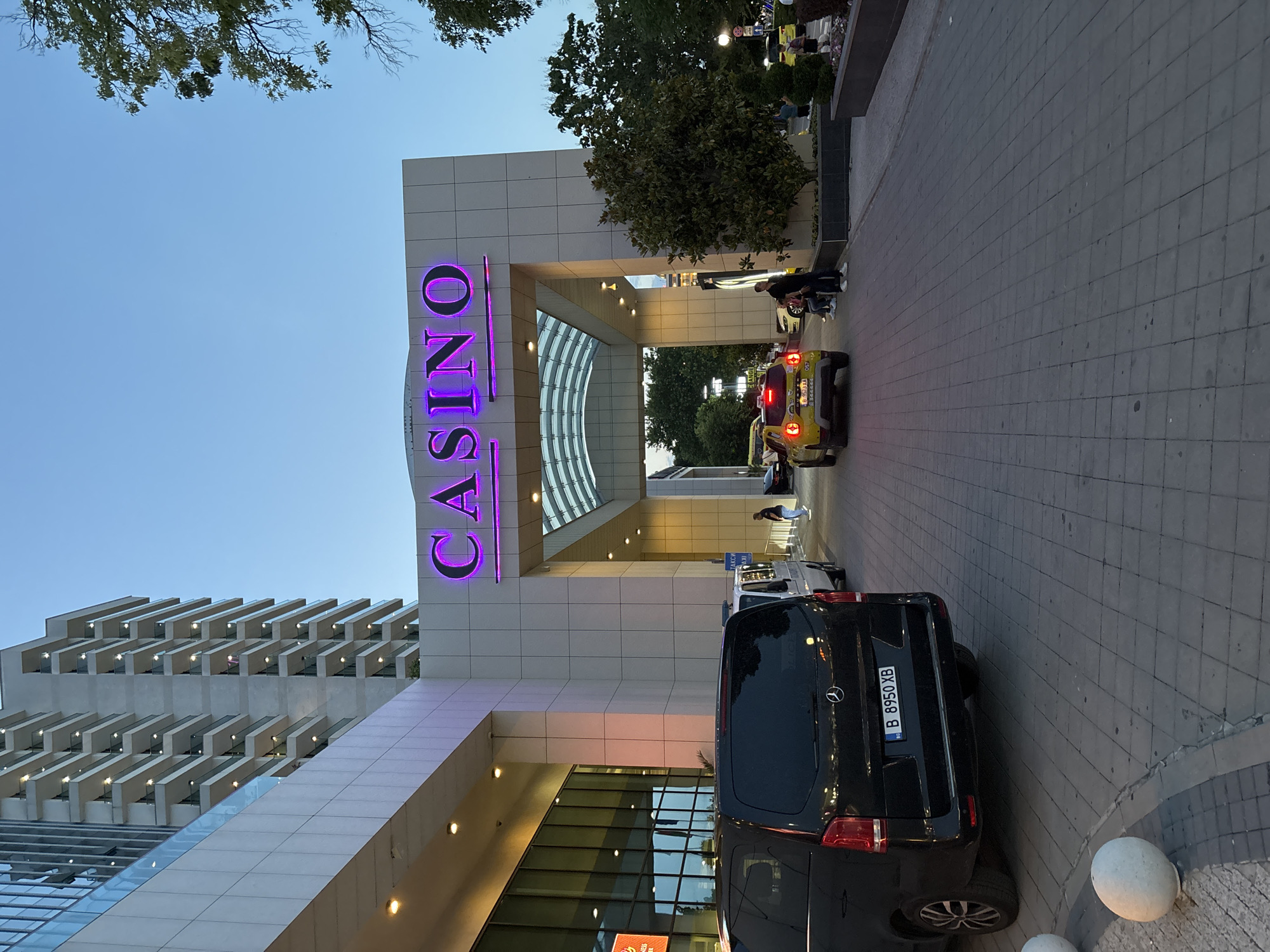 This image captures a modern building complex at dusk, featuring a prominent casino entrance. The scene is identified as being in Varna, Bulgaria. The central focus is a large, light-colored, multi-story structure with a contemporary design, characterized by an extensive glass facade on the left section and numerous open-air balconies on the upper residential or hotel floors. A grand, arched portico forms the main entrance, above which a large, illuminated purple neon sign clearly spells "CASINO."
In the foreground, a black Mercedes-Benz van is parked with its rear facing the viewer, displaying a Bulgarian license plate "B 8950 XB," indicating registration in Sofia. Further down the paved drive, a yellow Dacia Duster taxi is visible, also facing away, with a Bulgarian license plate "B 9573 XA," also registered in Sofia. Between the black van and the building, a white van, possibly a security or police vehicle, is partially visible. Various individuals are scattered across the scene: a group comprising two adults and two children stands near the yellow taxi, while other figures are seen walking or standing near the casino entrance and other vehicles.
The ground consists of grey interlocking pavers, with several white spherical bollards lining the edge of the paved area. Green trees and shrubs are situated to the right of the driveway and adjacent to the building. The sky above is a clear, transitioning twilight blue. Artificial lighting illuminates the "CASINO" sign, the building's facade, and interior spaces, with vehicle taillights also active. A blue sign with "TAXI" written in Cyrillic is visible in the background, and a faint "BUSINESS CENTER" sign can be discerned through the glass facade on the left.