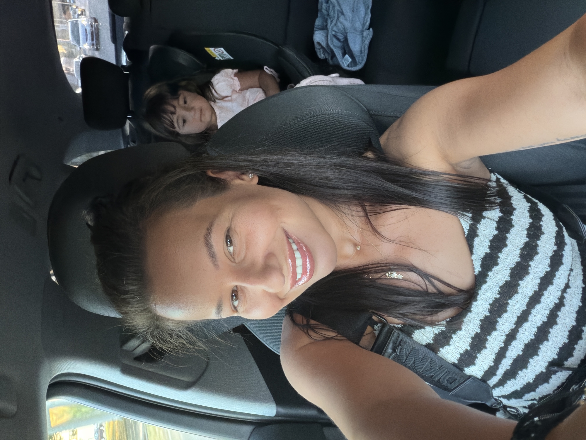 This media file is a selfie taken inside a car. The primary subject is a woman smiling at the camera. She has long, dark hair and is wearing a black and white striped top. A young girl is visible in the background, seated in a car seat, appearing to be asleep or resting. She is wearing a light pink outfit. The setting is the interior of a car, with car seats and the vehicle's interior visible. The time of day appears to be daytime, indicated by the natural light filtering into the car. The overall mood is relaxed and familial. There is no visible text in the image. The location context is Burgas, Bulgaria.